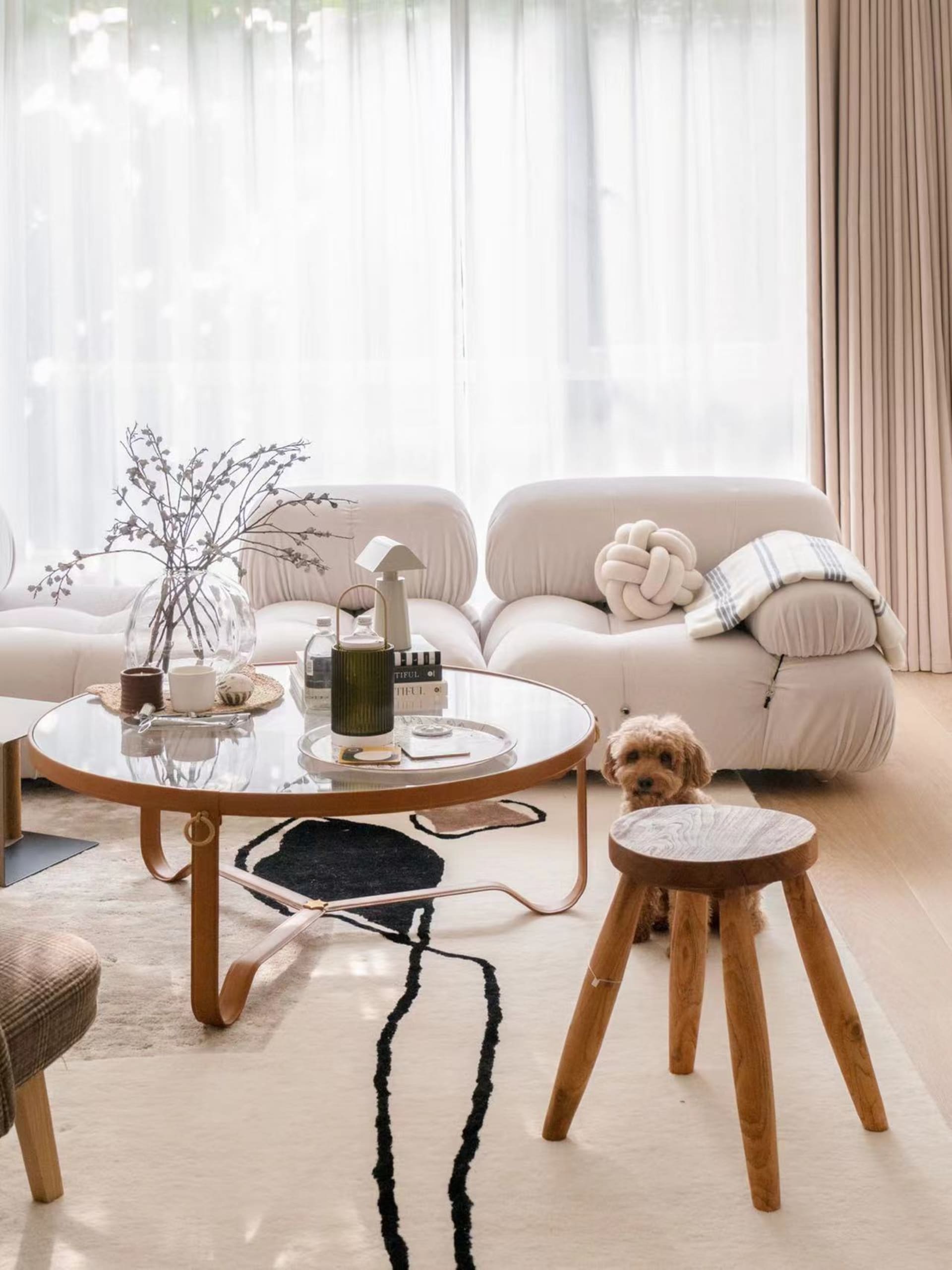 A cozy, modern minimalist living room with white textured fabric furniture, a wooden stool, and a glass coffee table adorned