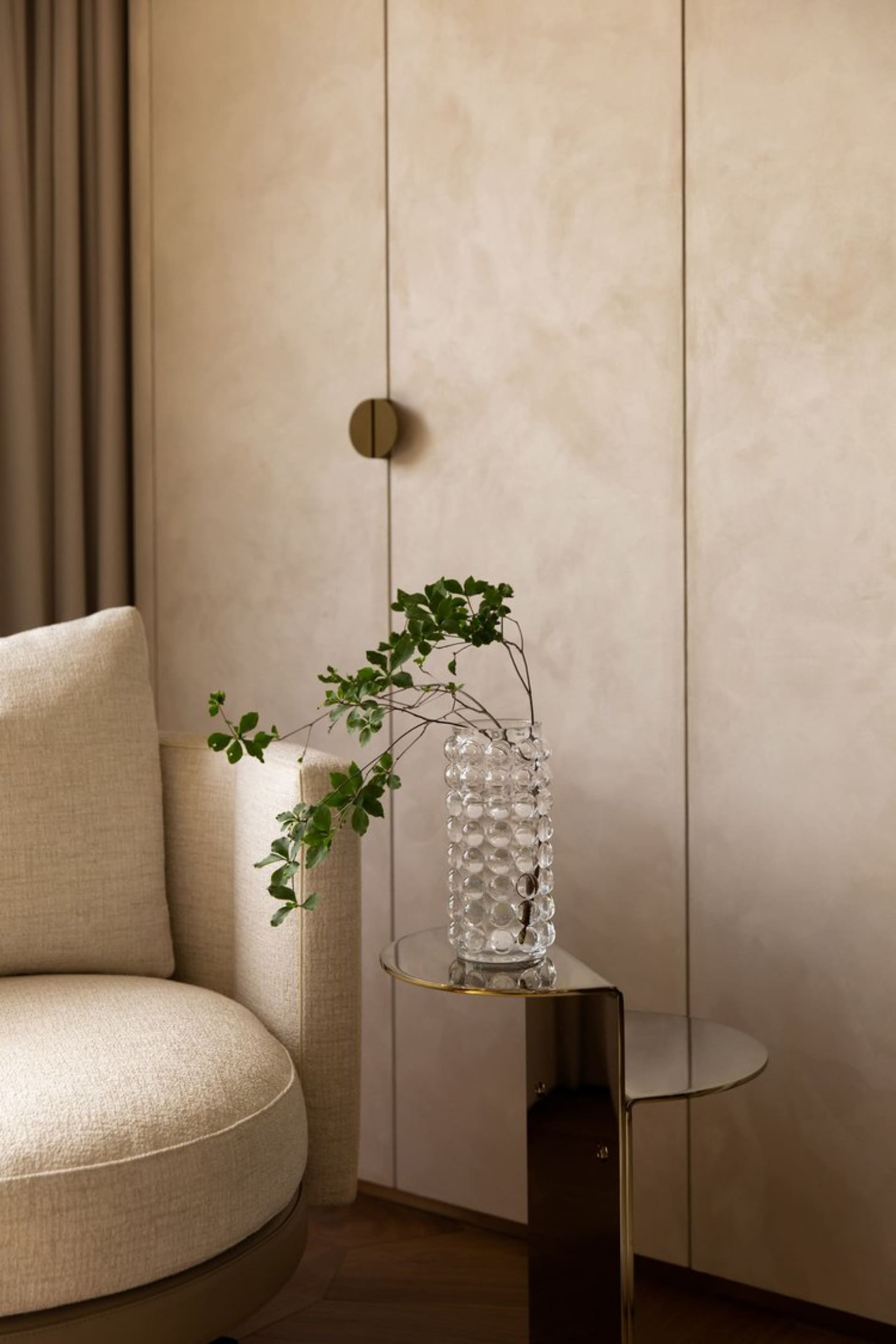 Modern minimalist living room with glass vase and metallic side table against a textured beige wall.