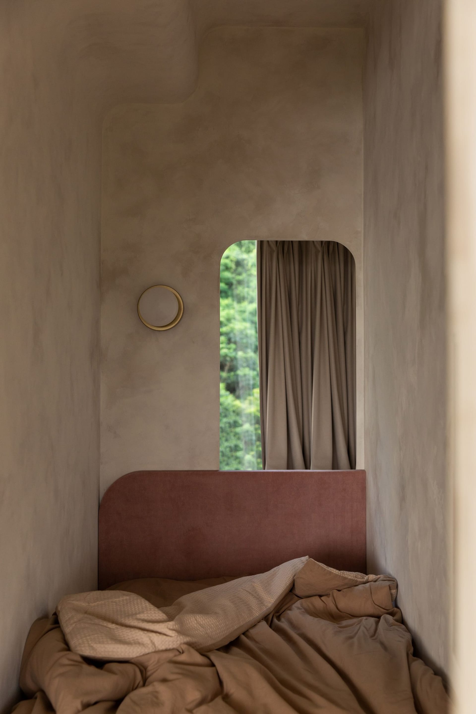 A minimalist bedroom with a pink velvet headboard and a cozy atmosphere, featuring a round light on the wall and a view of gr