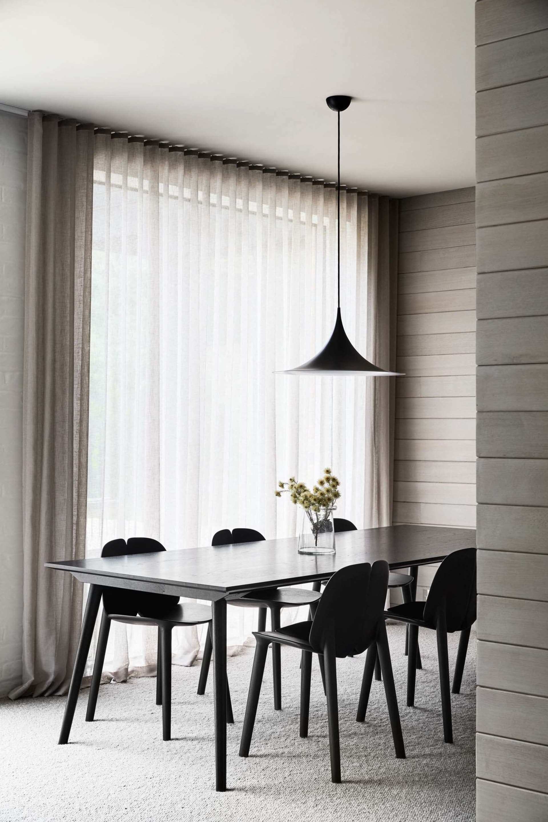 A minimalist dining room featuring a long wooden table with black chairs, large windows with sheer curtains, and a black pend