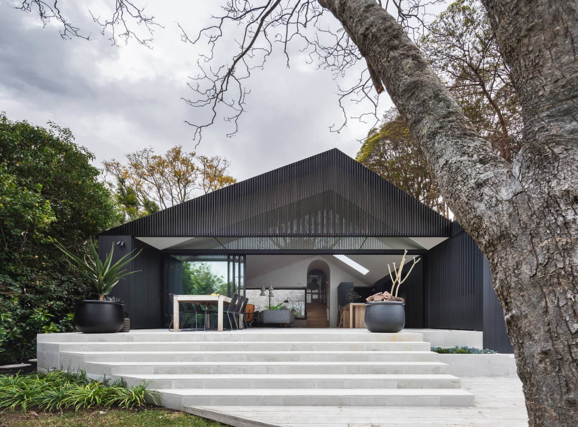 A modern residential exterior with black corrugated metal siding, large glass windows, and a large tree in the foreground.