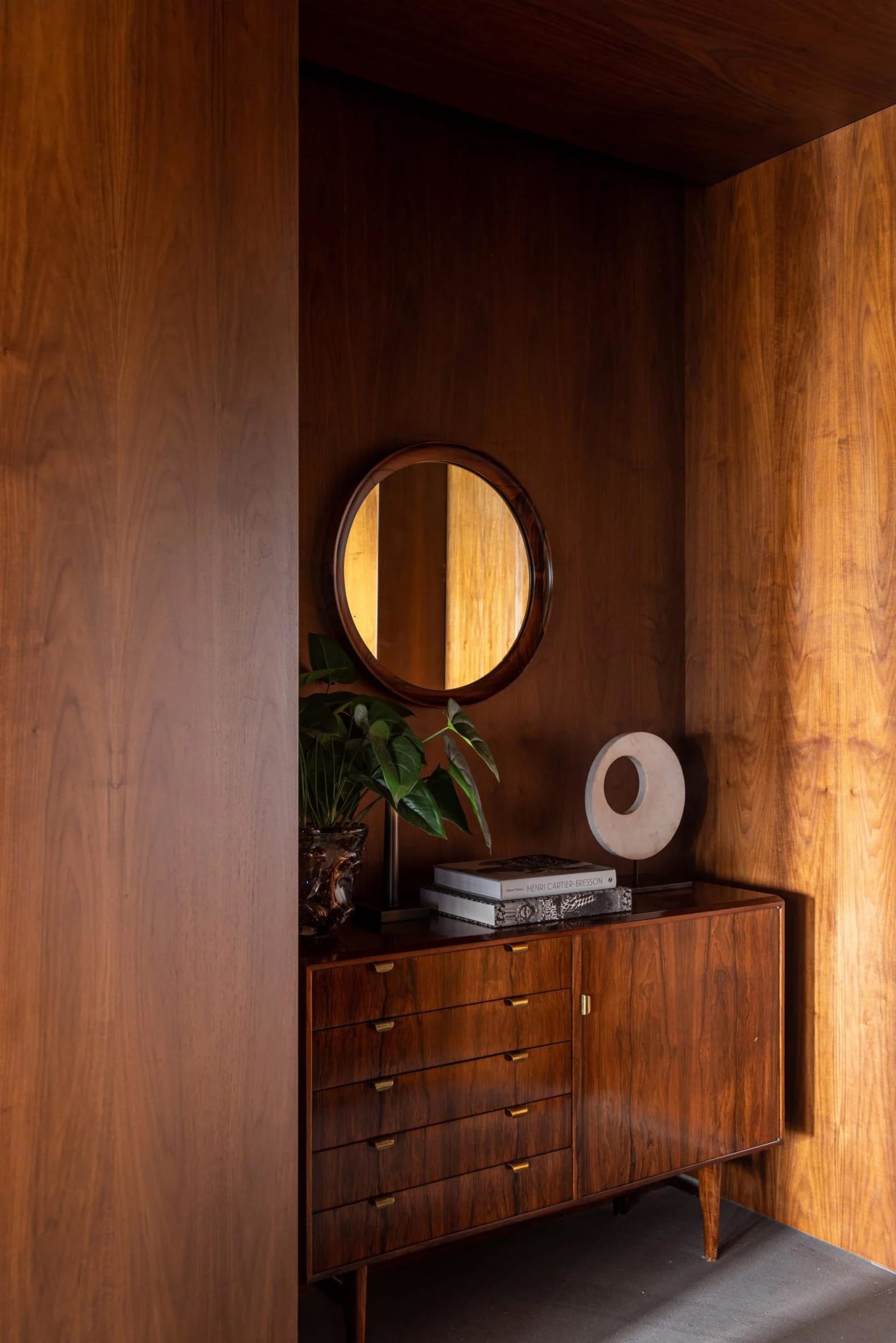 A modern home library featuring minimalist wooden furniture, a round wooden mirror, a potted plant, and a stack of books.