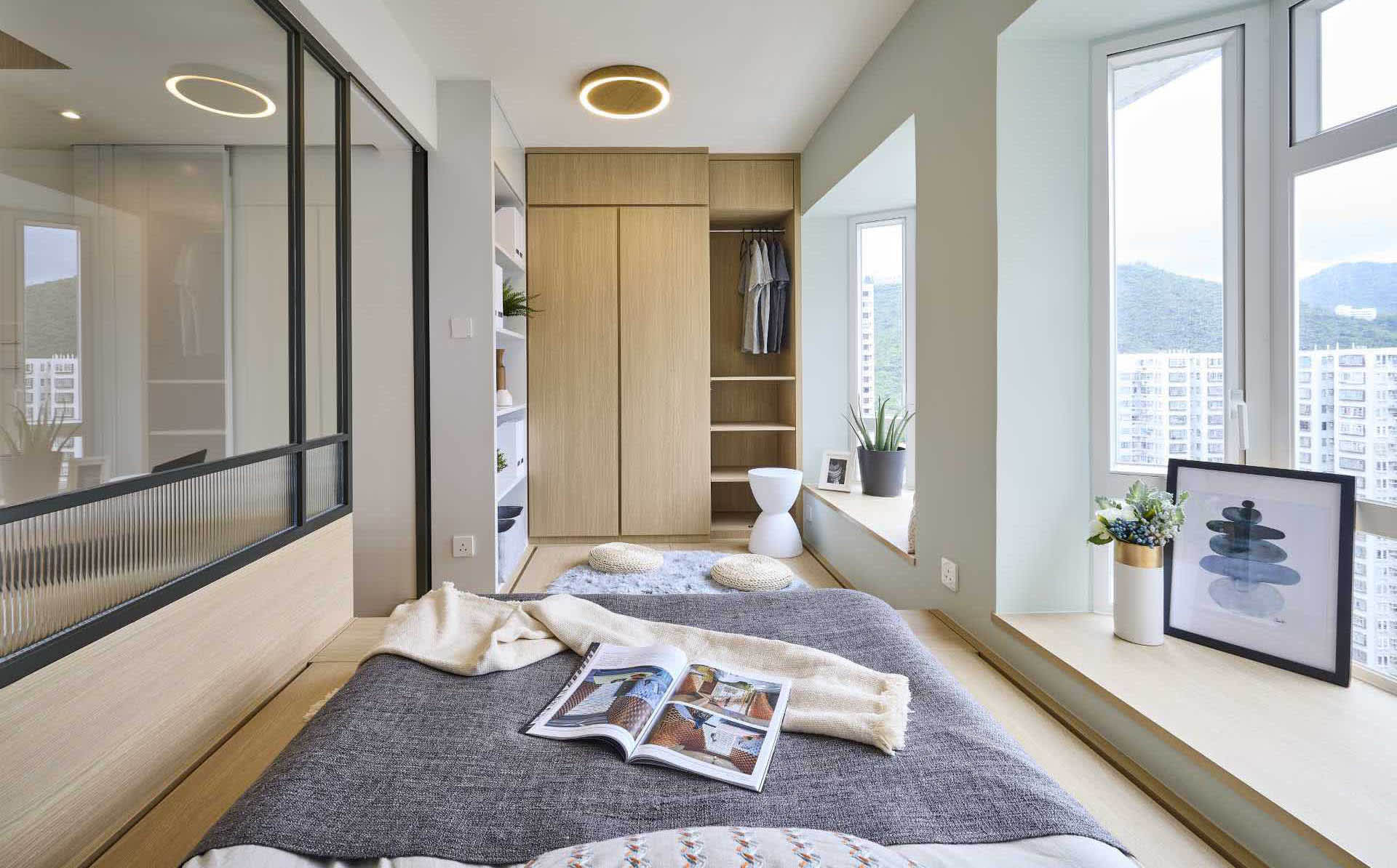 A modern minimalist bedroom features a bed with a gray bedspread and a magazine lying on it, a wooden wardrobe, and a window