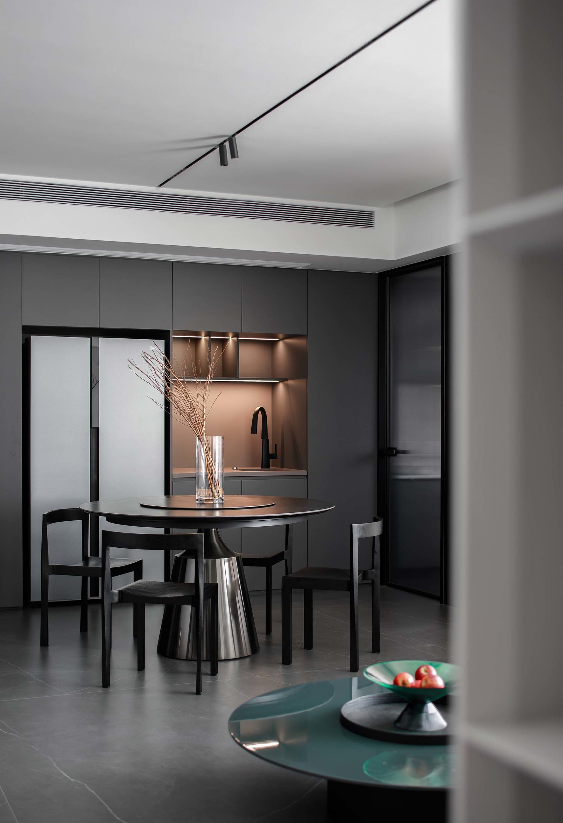 A modern dining room features a metallic table with dark chairs, illuminated by sleek lighting and surrounded by minimalist b