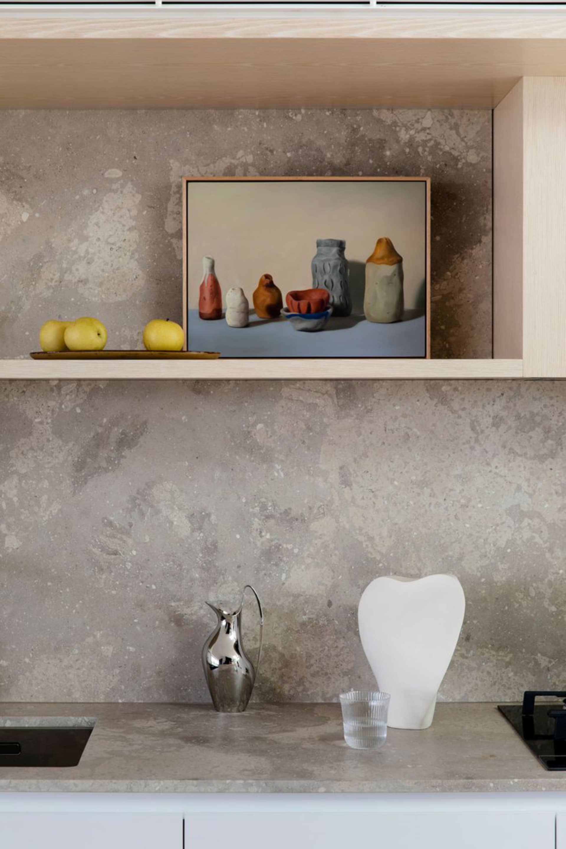 A modern kitchen counter with a grey concrete finish, featuring a metal pitcher, a white vase, and an artwork of vases and ap