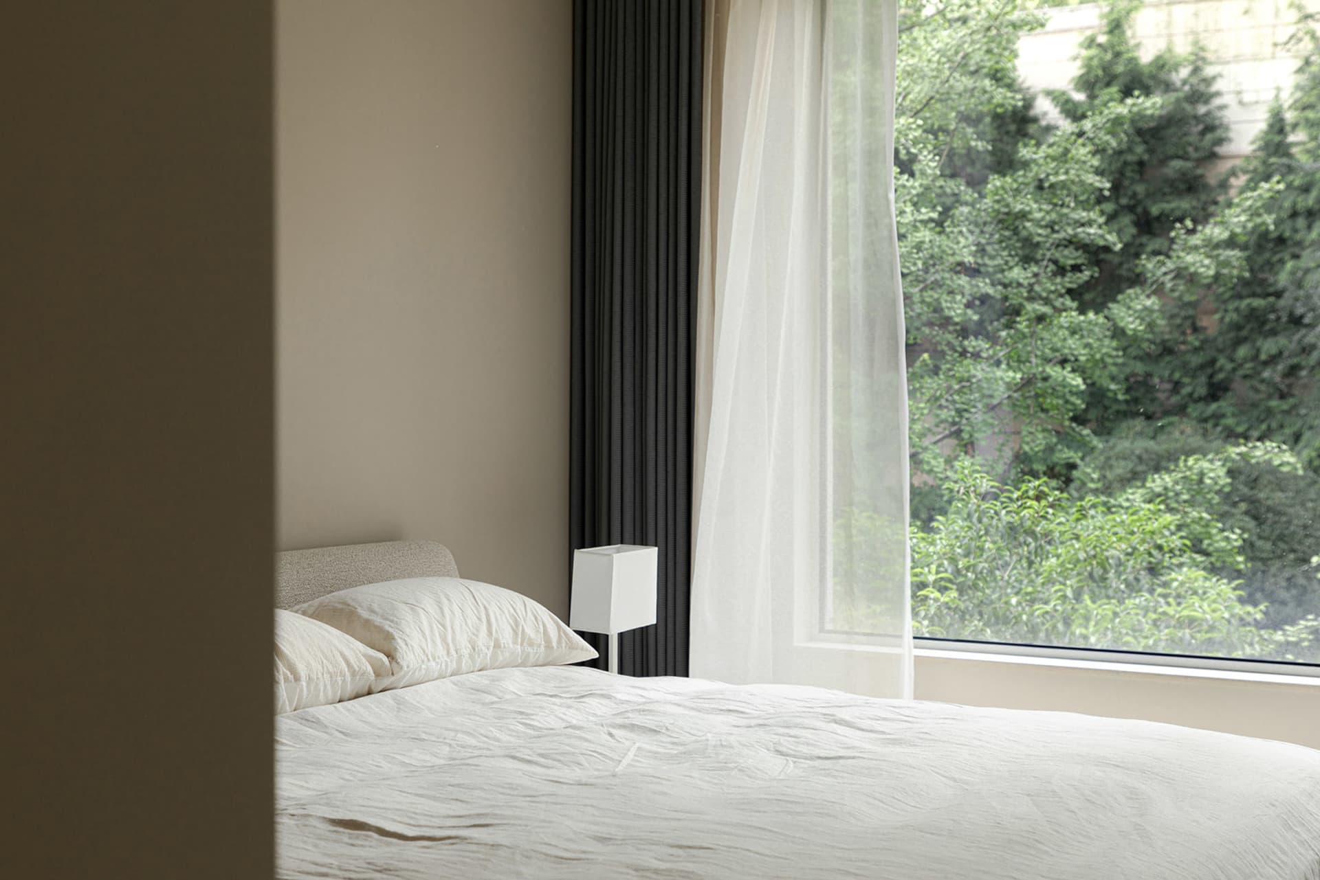 A minimalist bedroom with a bed featuring white textiles and natural light streaming through a large window with a sheer curt