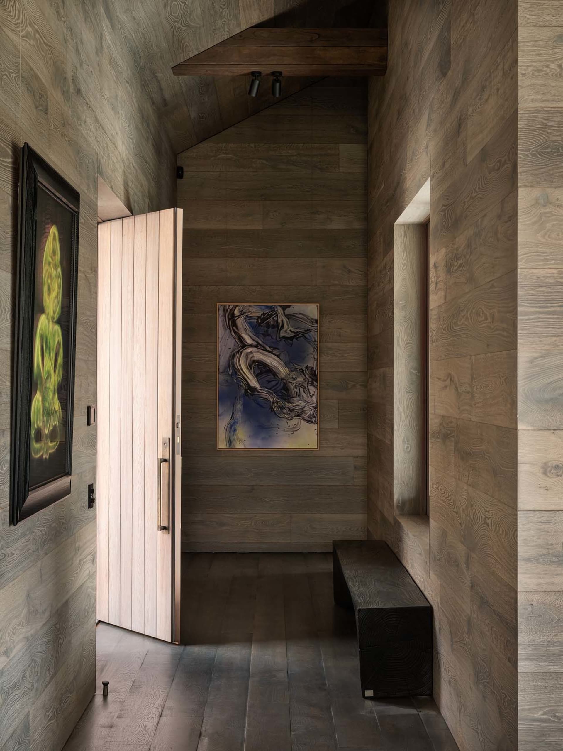 A modern entryway featuring wooden walls and a door, with abstract artwork and a bench.
