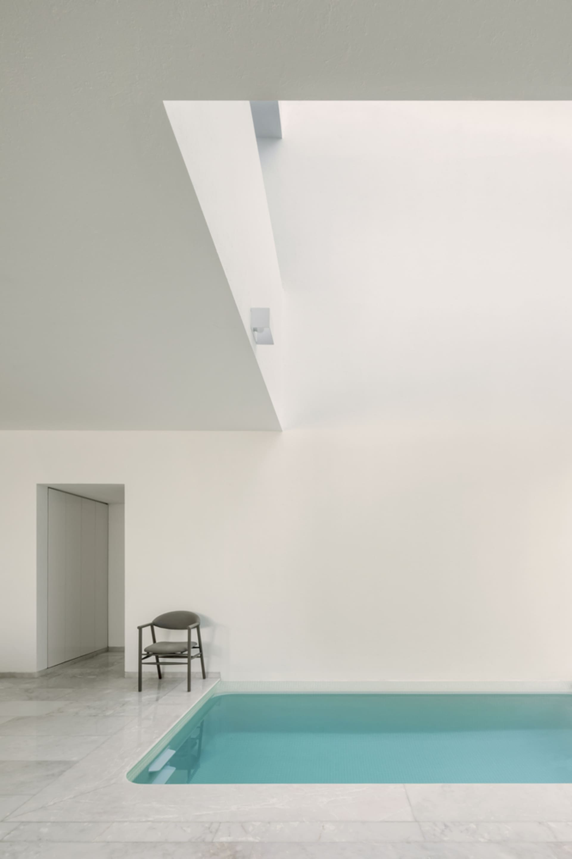 A minimalist indoor swimming pool with a white marble floor, surrounded by white walls and a ceiling with a skylight.