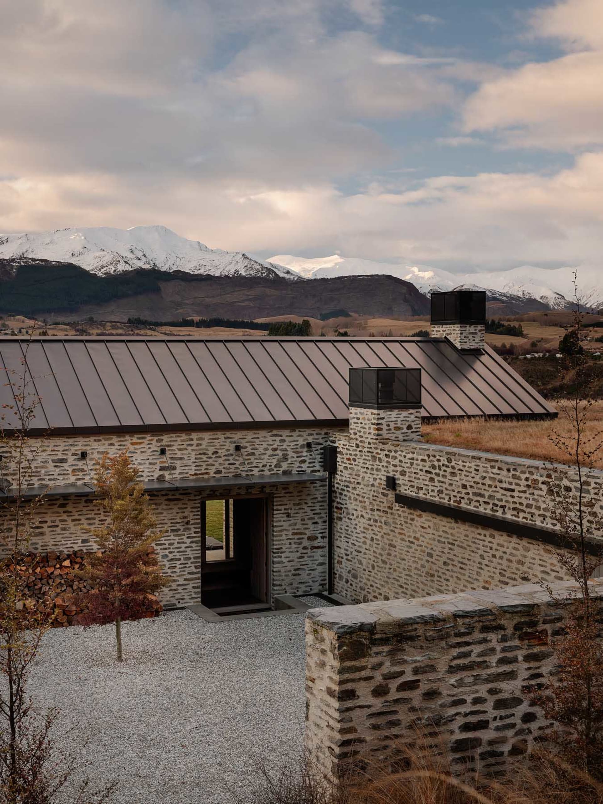 A stone cottage with a metal roof situated on a gravel driveway with a backdrop of snow-capped mountains.