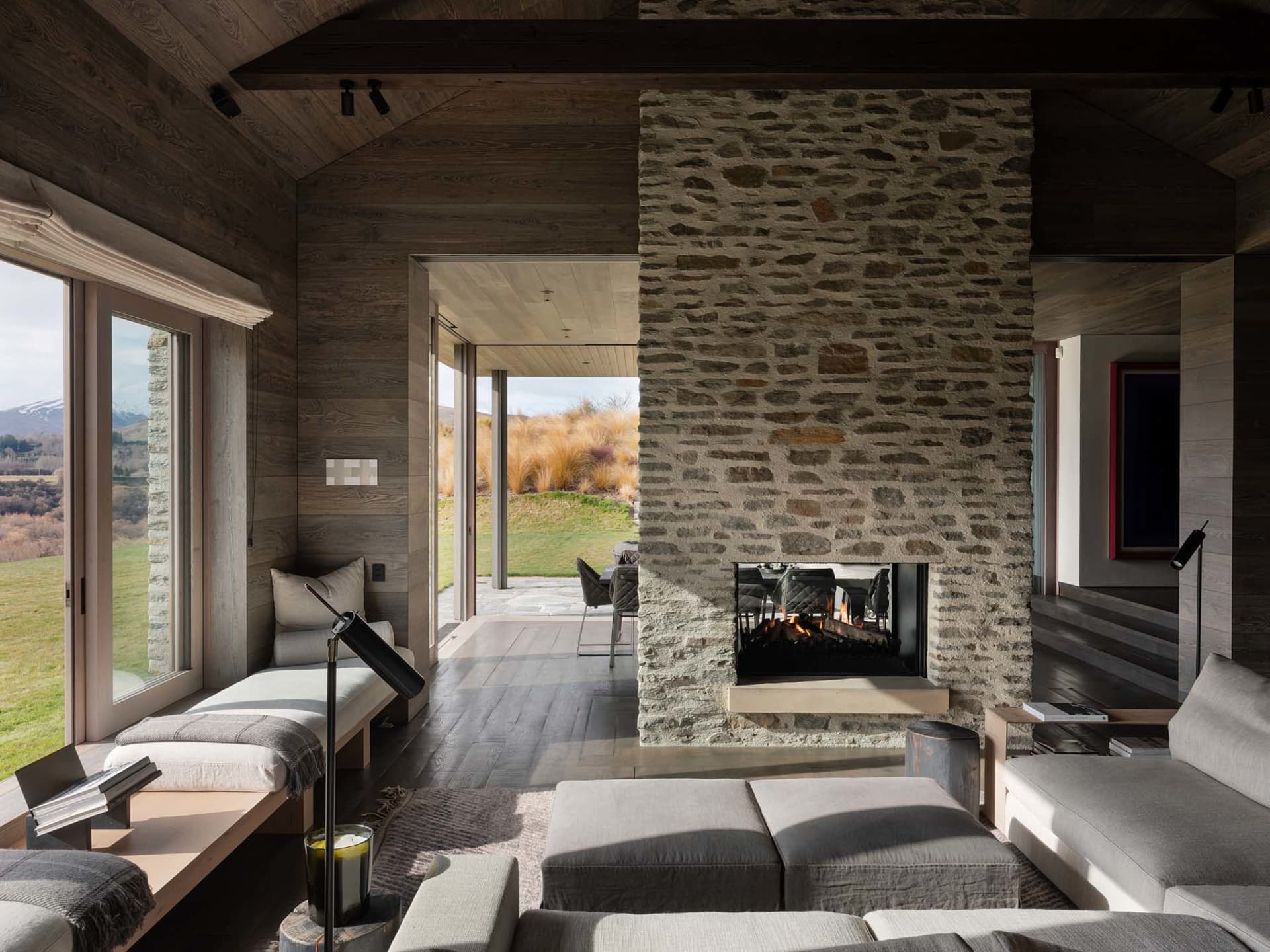 A cozy modern rustic living room with stone fireplace, wooden beams, and large windows overlooking a scenic landscape.