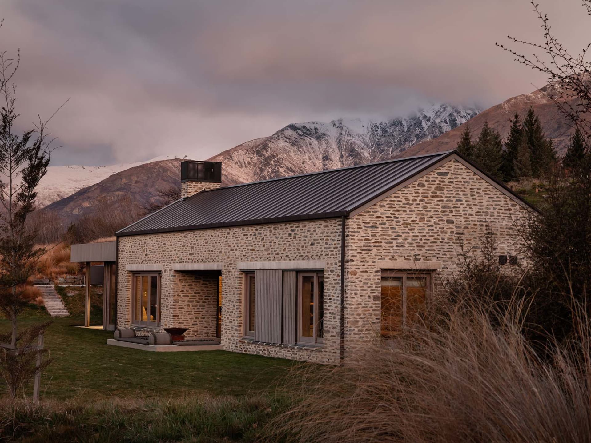 A rustic stone residential exterior with a metal roof and a stone chimney, set against a mountainous backdrop.