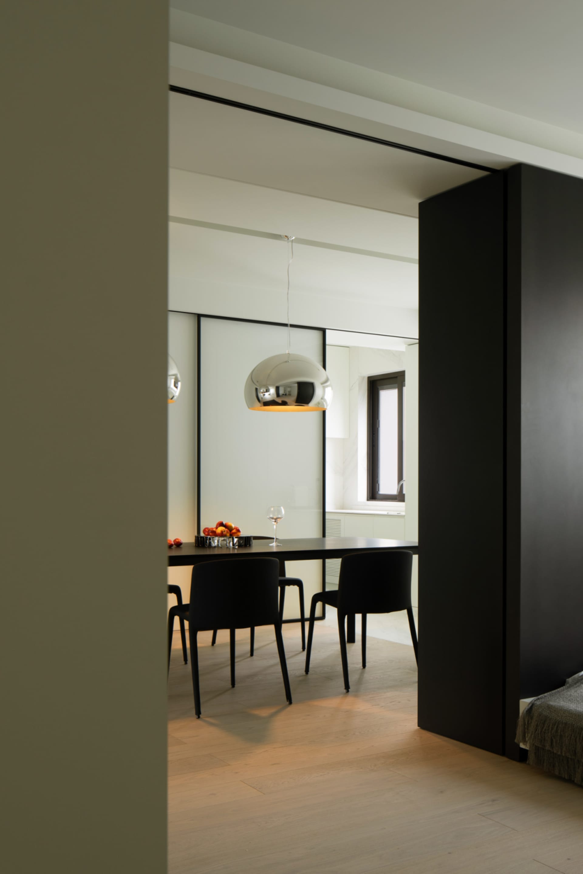 A modern minimalist dining room featuring a metallic pendant light fixture, black chairs, and a wooden floor.