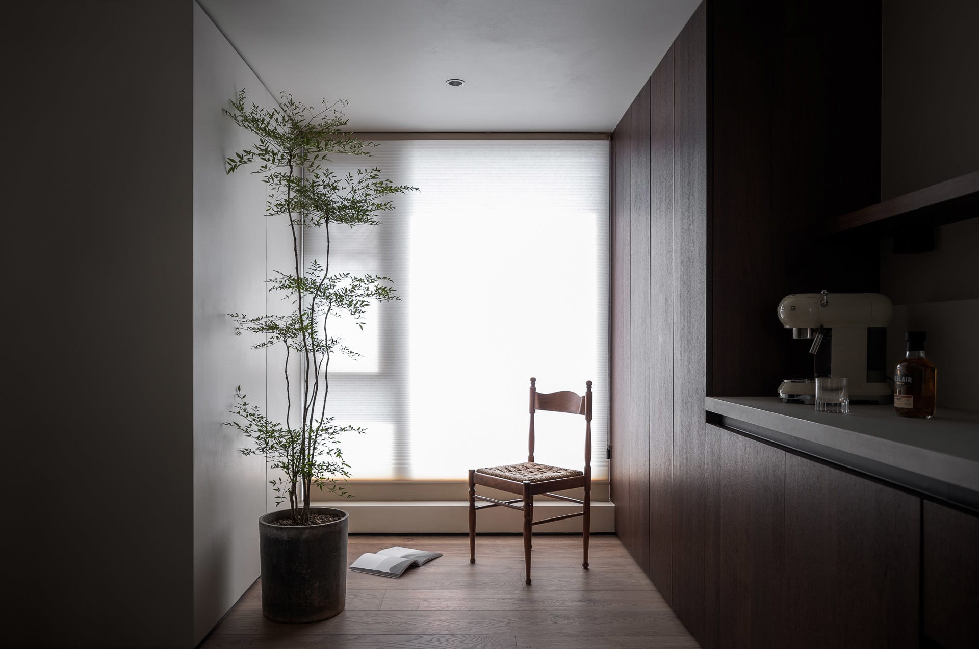 Minimalist home office space featuring wooden chair, potted plant, and glass window with blinds.