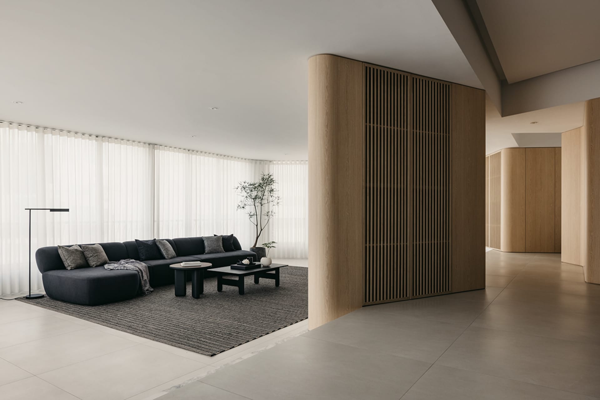 Modern minimalist living room with a black sectional sofa, wooden paneling, and a large floor lamp. Floor-to-ceiling windows