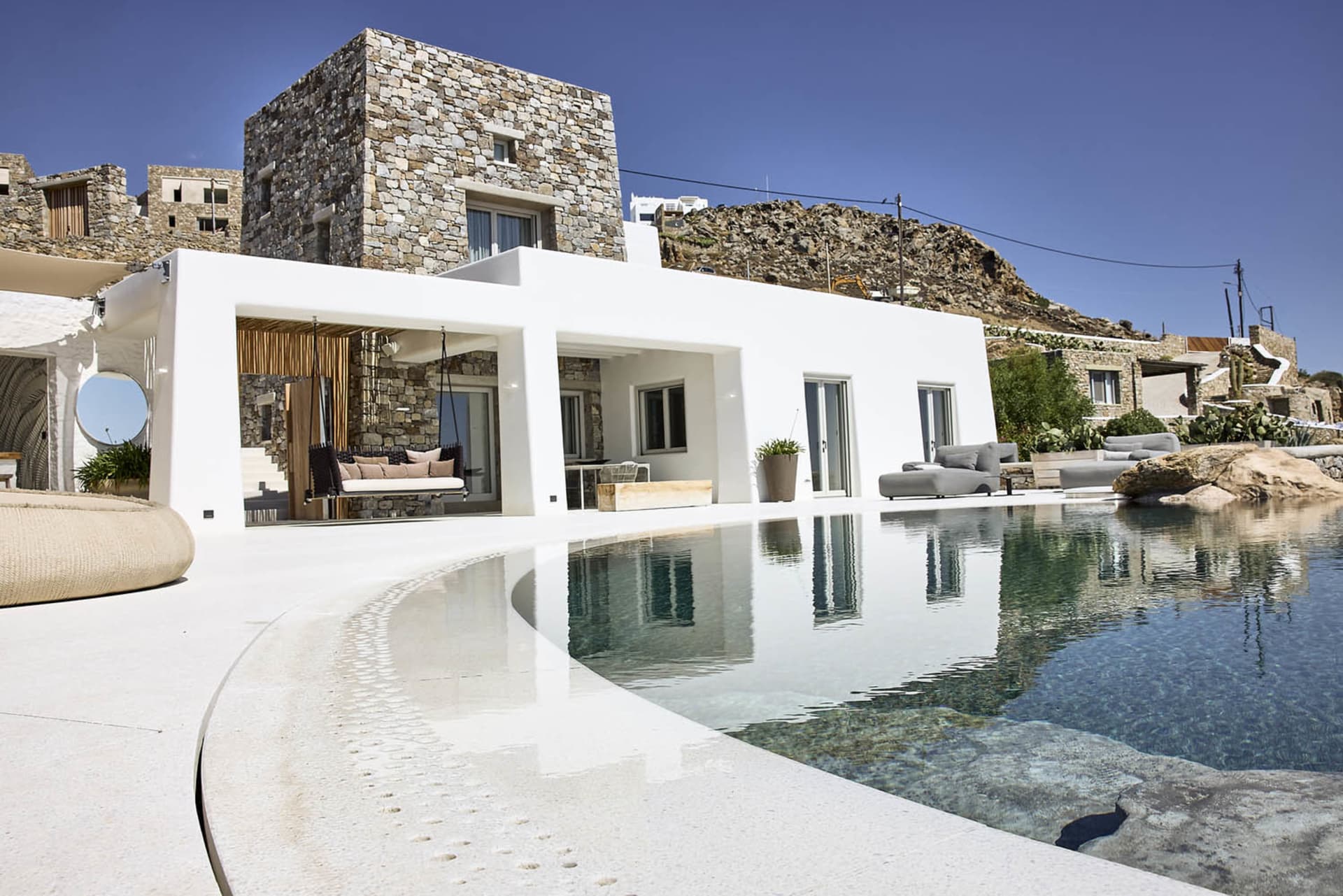 A minimalist residential exterior featuring white plaster and stone materials, situated on a hillside with an infinity pool.