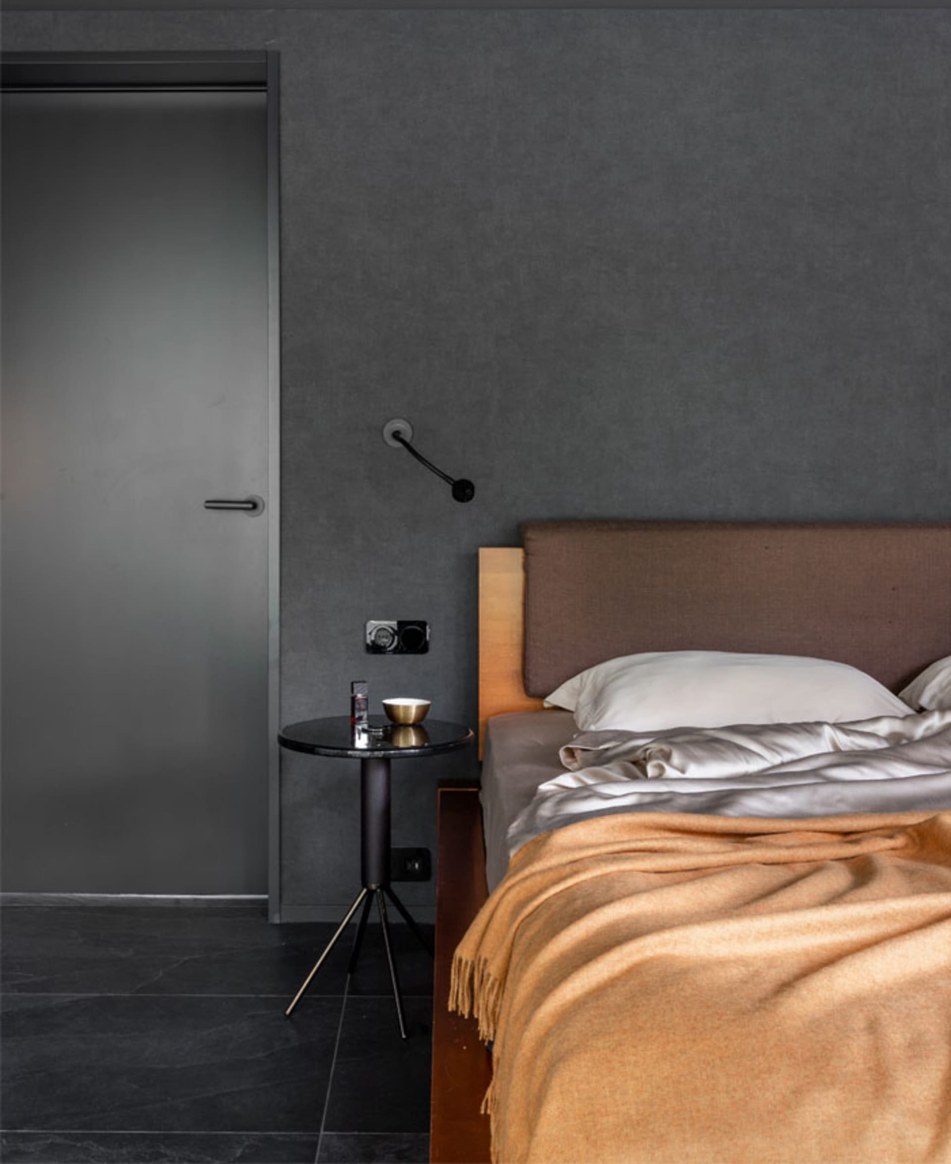 A minimalist bedroom featuring a bed with a brown upholstered headboard, a yellow blanket, and a black side table with a lamp