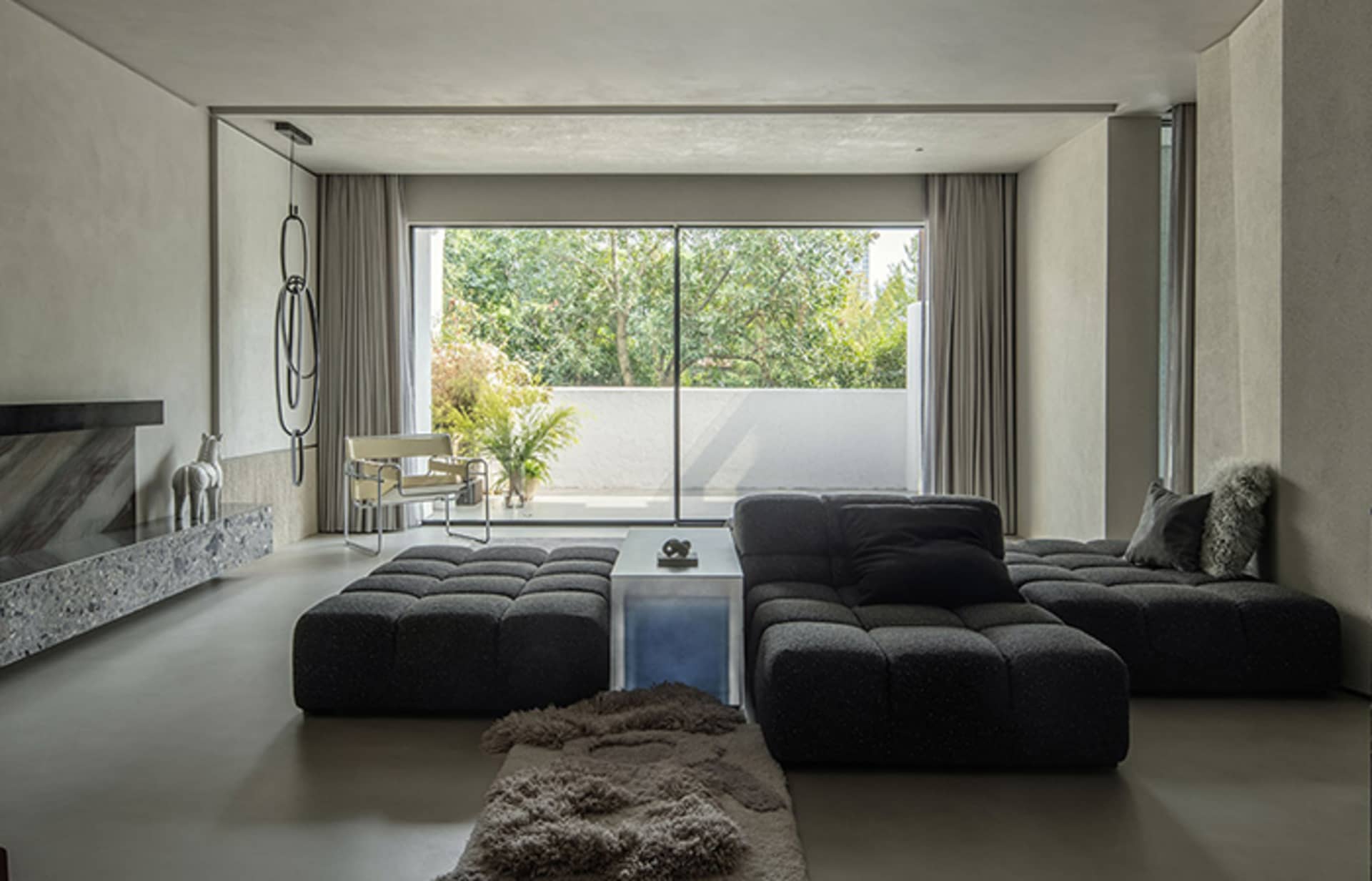A modern minimalist living room with a concrete floor, large windows, and a large black modular sofa.