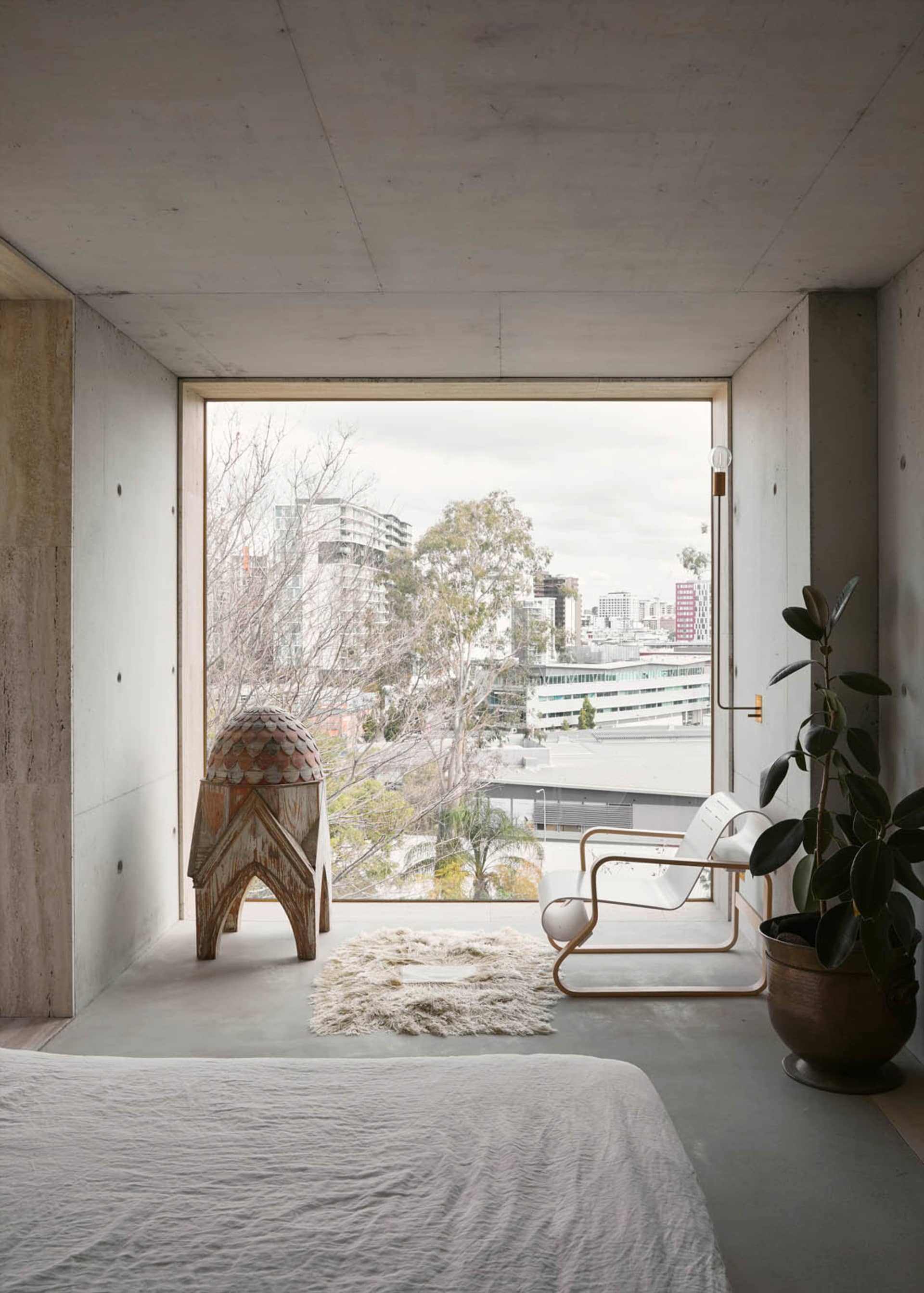 A minimalist bedroom with concrete walls, a white bed, a white chair, and a plant in a large window with a city view.