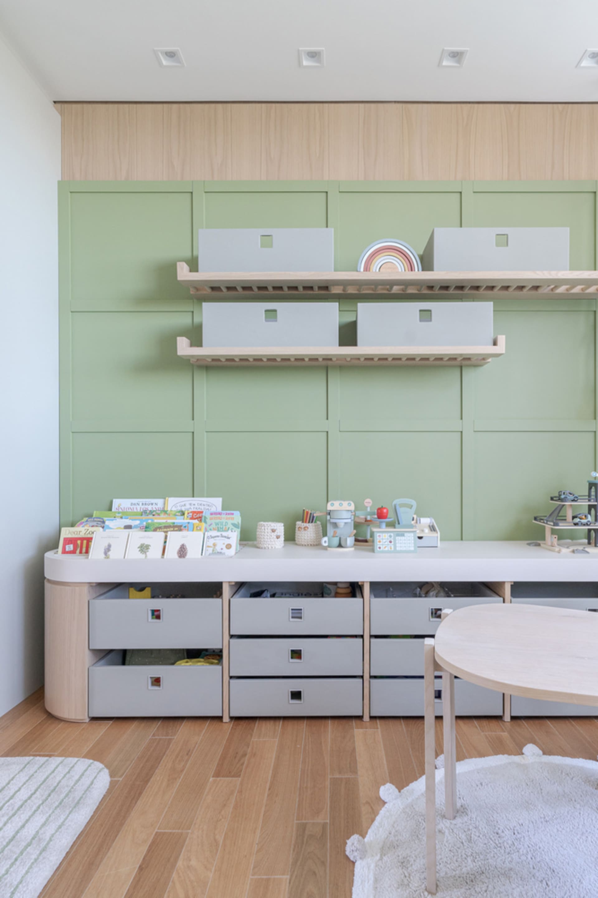 A modern playroom with green shelves, wooden floor, and white furniture, featuring toys and books.