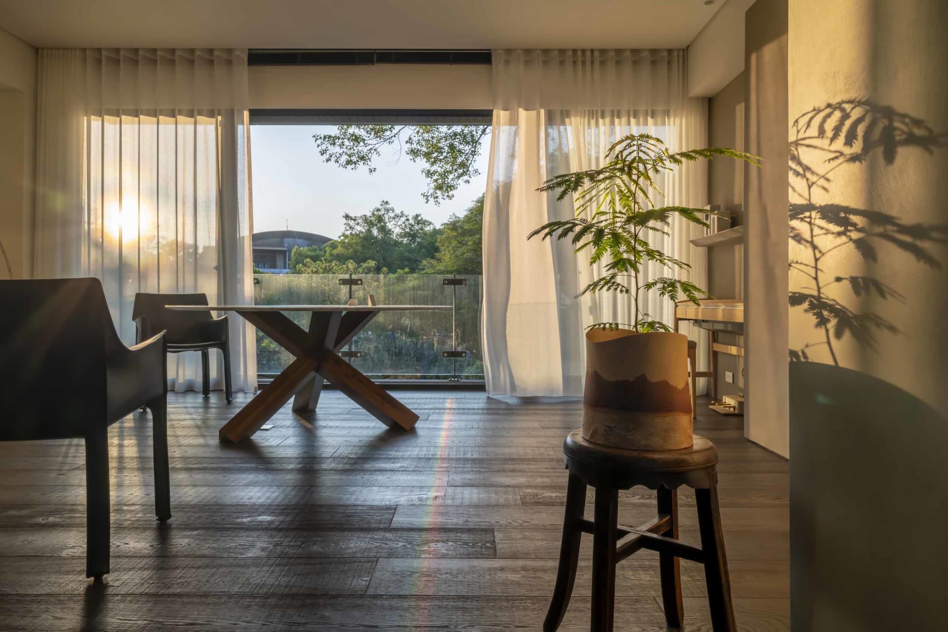 A contemporary residential interior featuring wooden flooring, large windows with a view of greenery, and a minimalist design