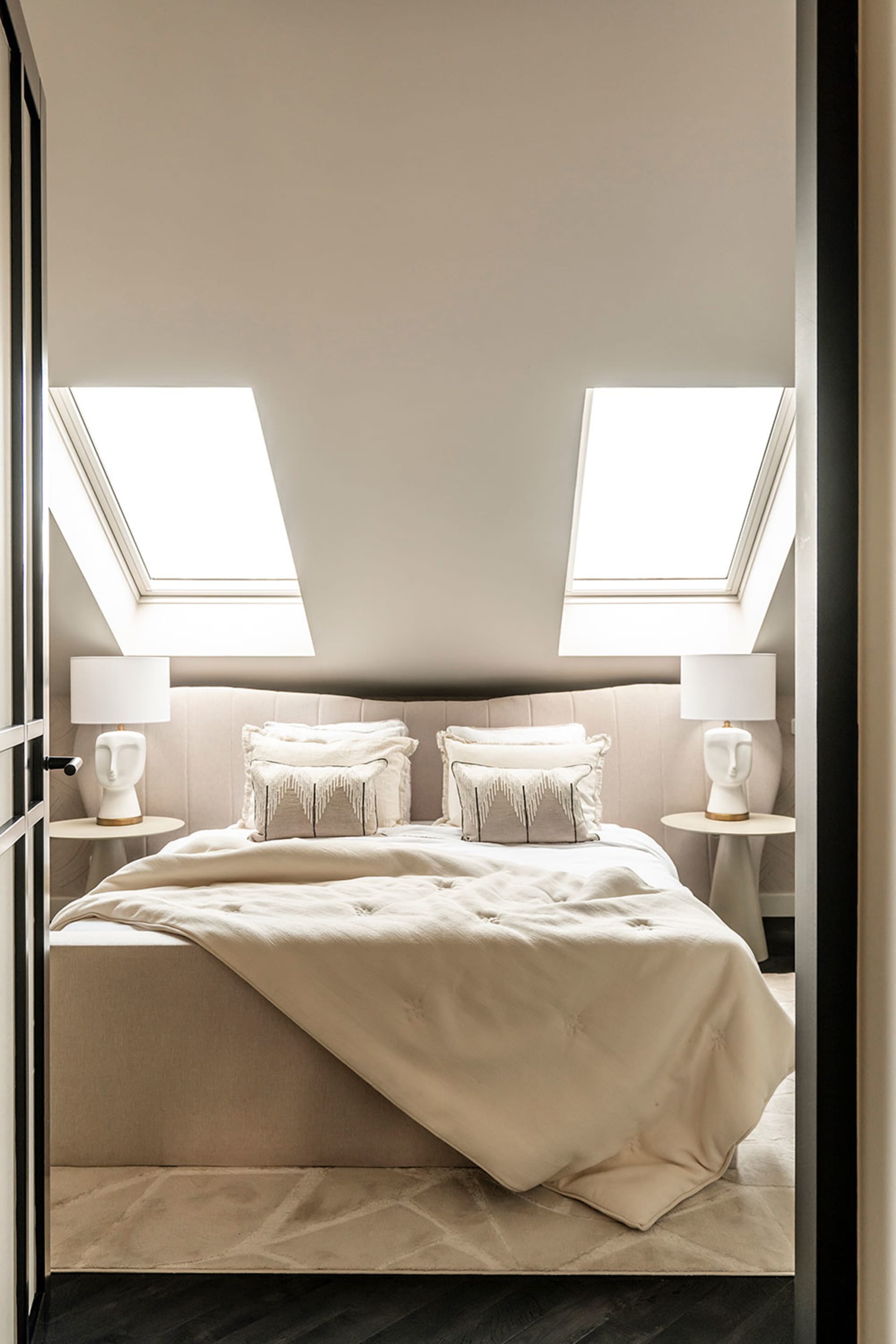 Modern, minimalist bedroom with white bedding, pillows, and a light-colored fabric headboard, featuring skylights and a cozy