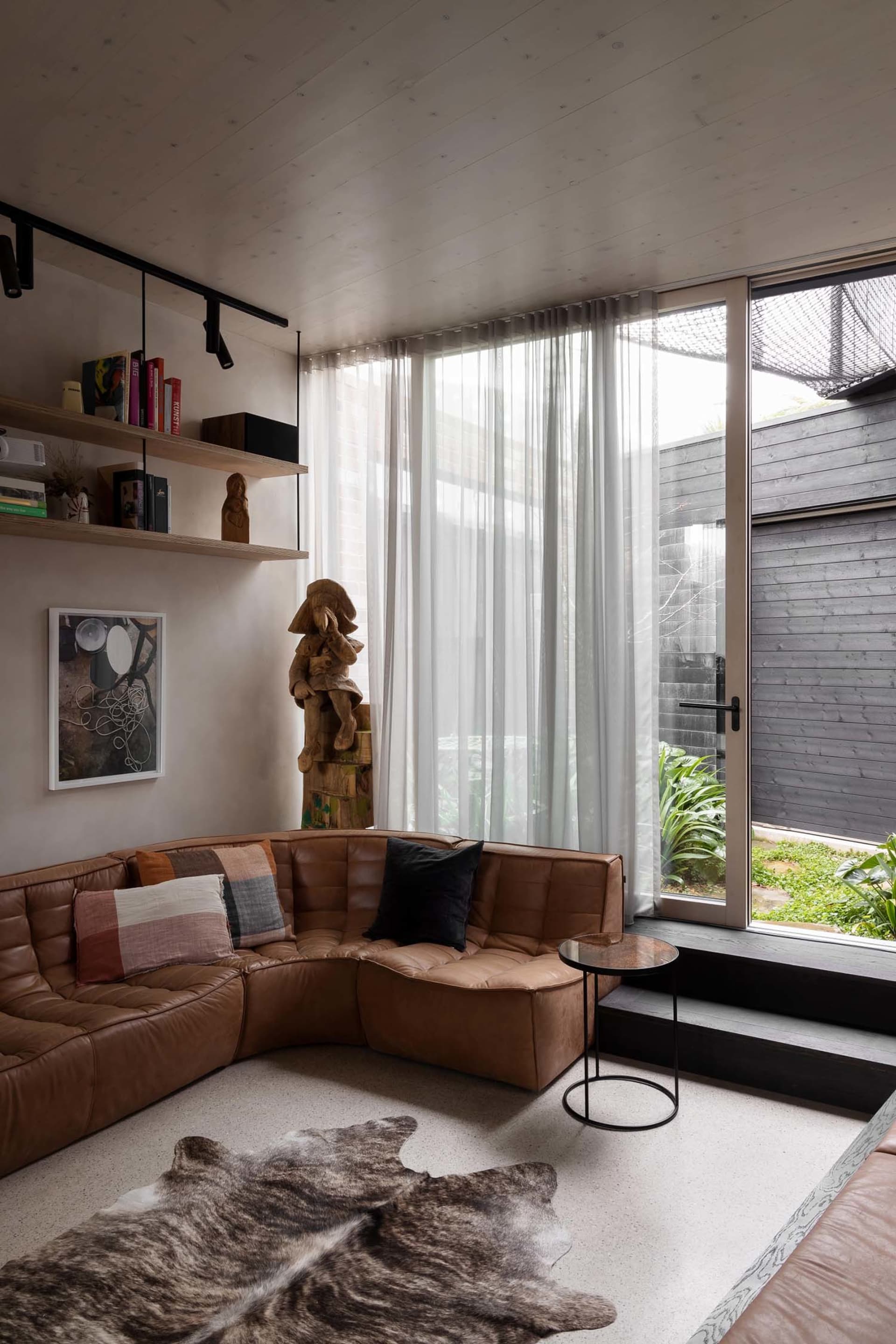 A living room with a brown leather sectional sofa, glass coffee table, wooden floor, and large windows with sheer curtains.