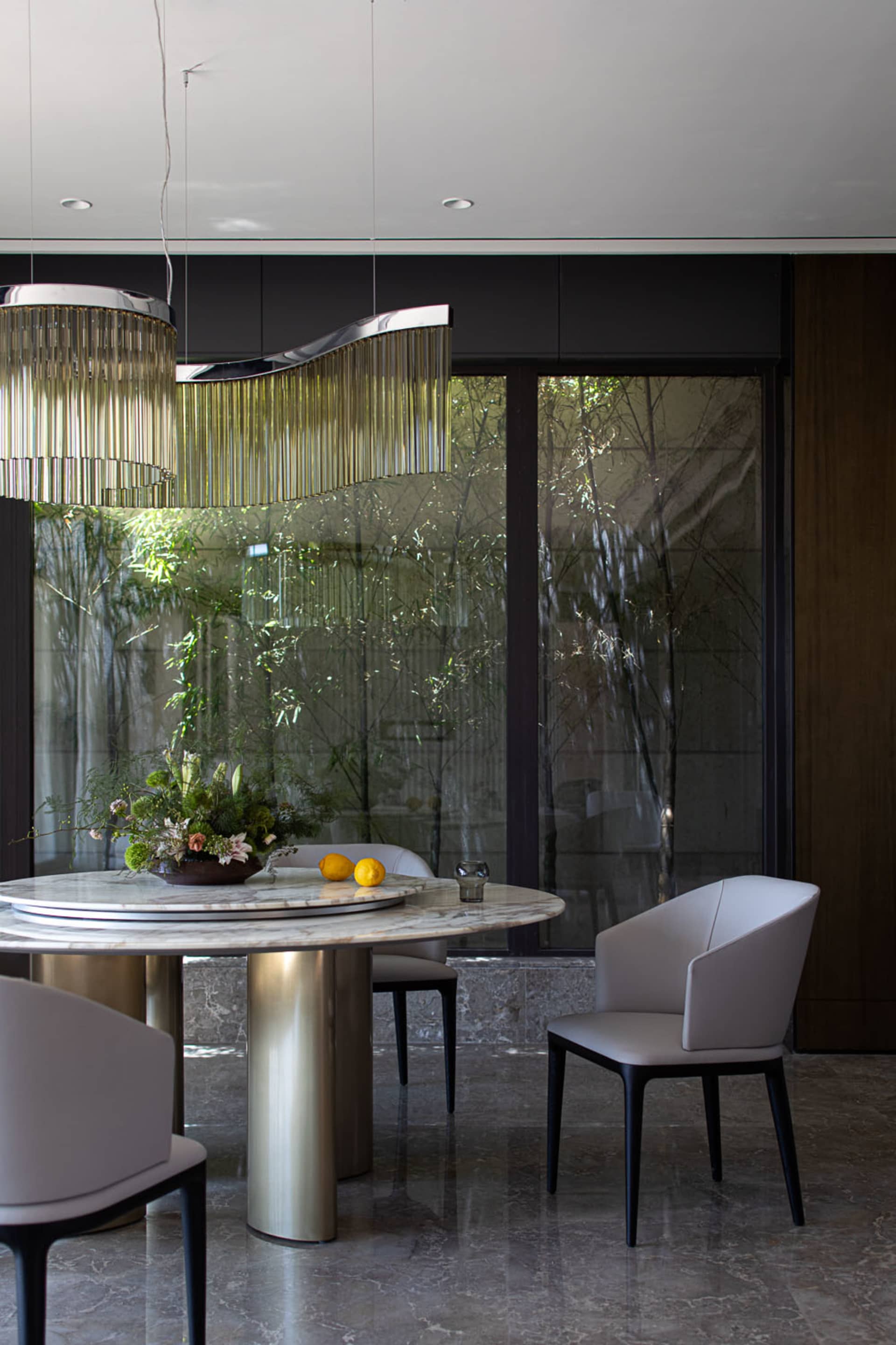 A modern dining room featuring a marble table and chairs, with a glass wall behind and a hanging chandelier.