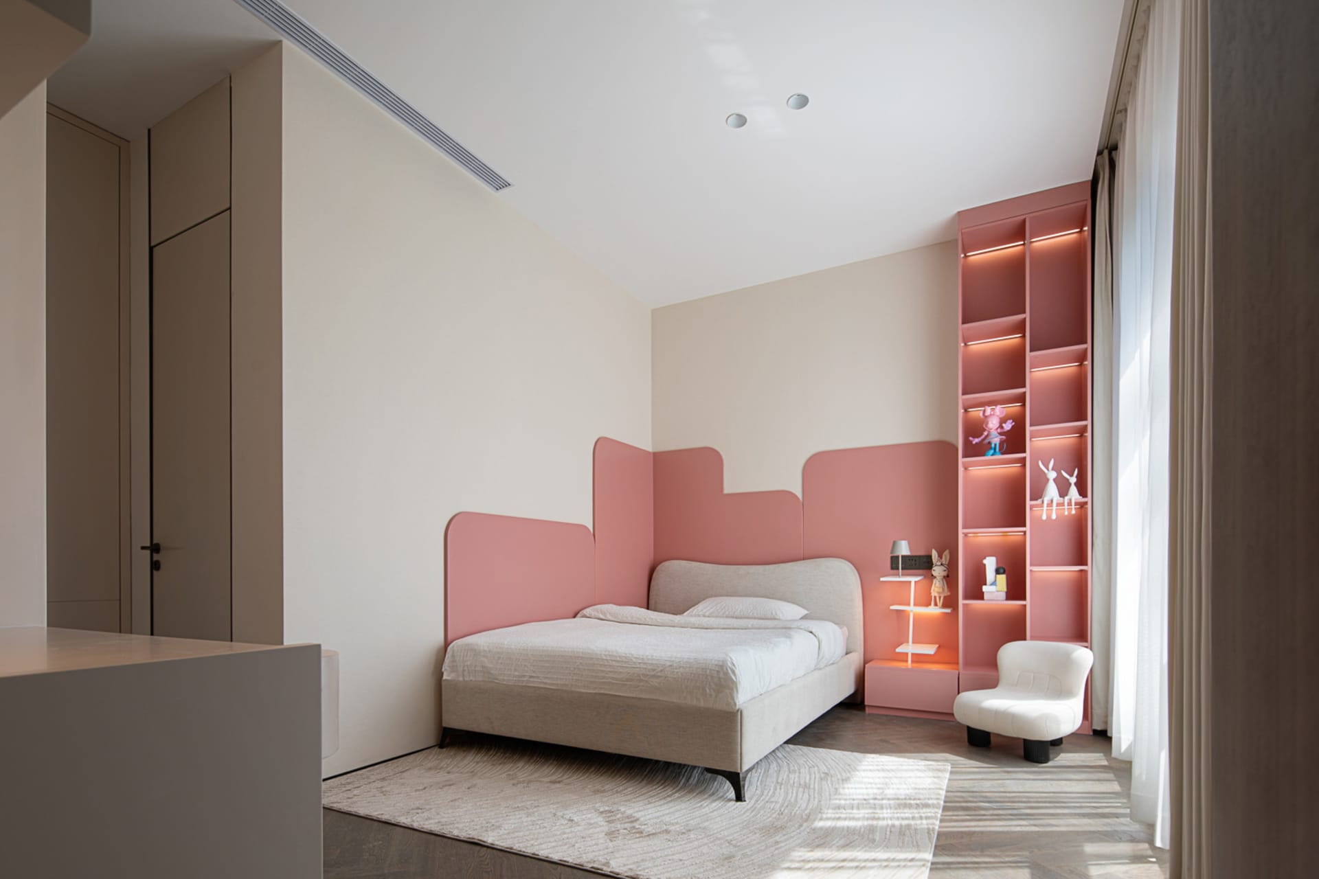 A modern bedroom featuring a pink bed, white chairs, a pink bookshelf illuminated by lights, and white curtains.