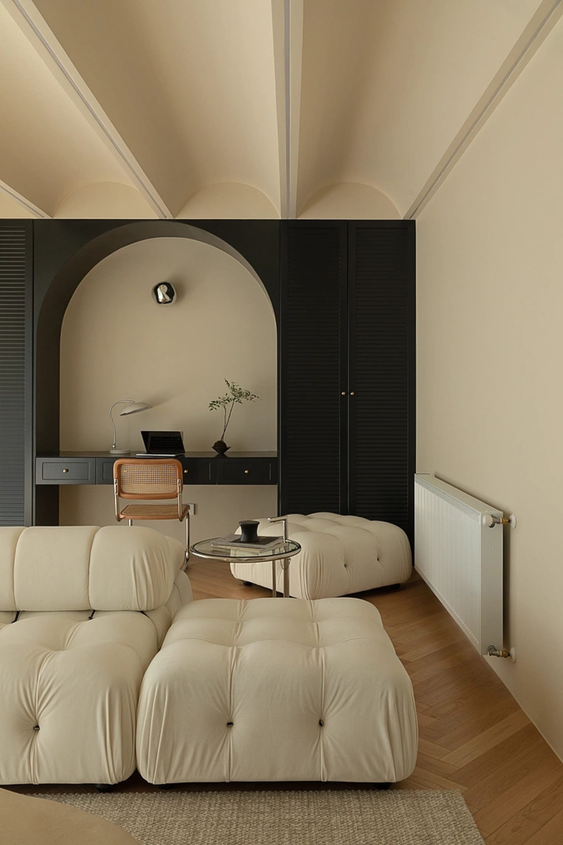 A minimalist living room with a white fabric sofa, ottoman, and a black desk with a chair, all set against a cream-colored wa