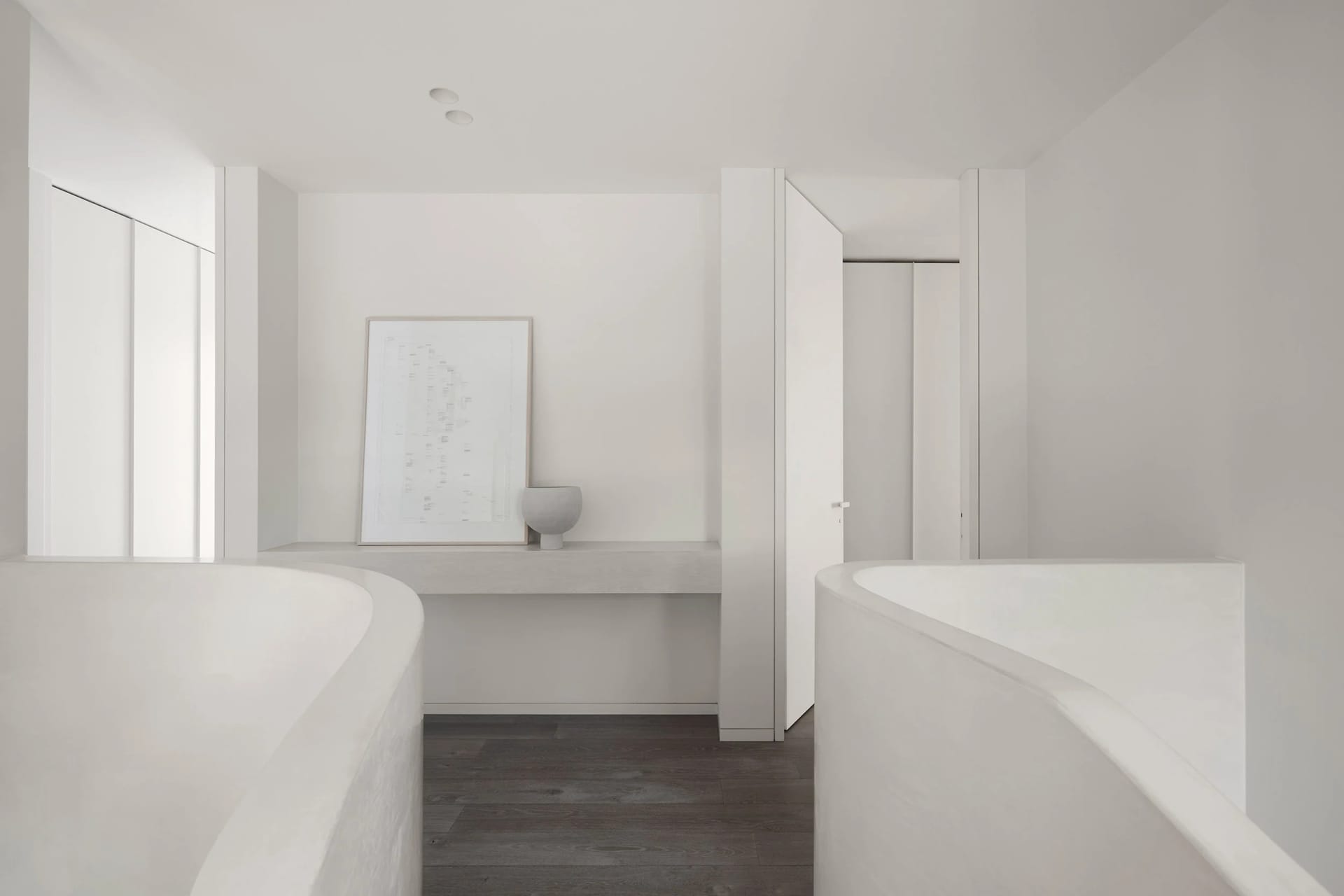 A minimalist bathroom with two concrete bathtubs, dark wooden flooring, and a small shelf with a vase and framed artwork.