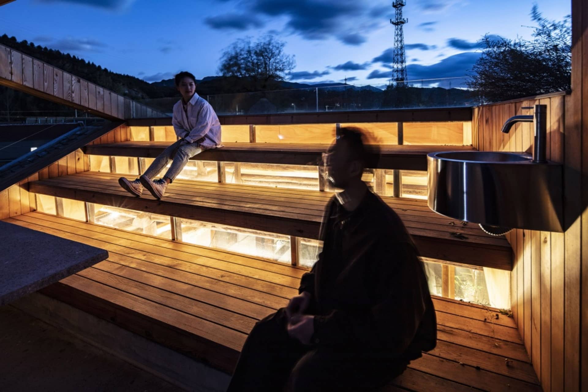 Two people sitting on wooden steps lit by warm lights during sunset.