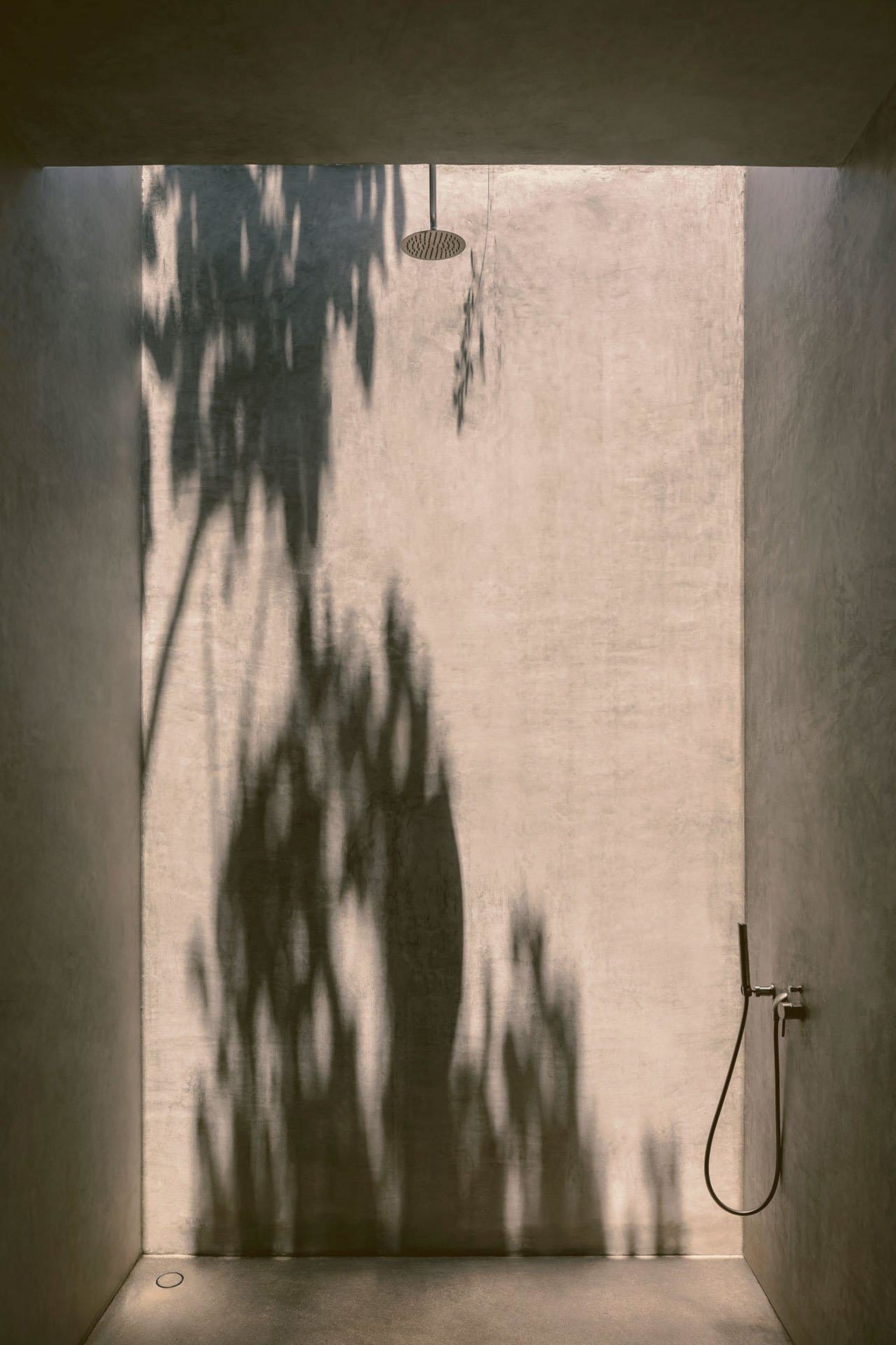A minimalist concrete bathroom features a circular shower head and a shadow of trees cast on the wall, creating a serene and