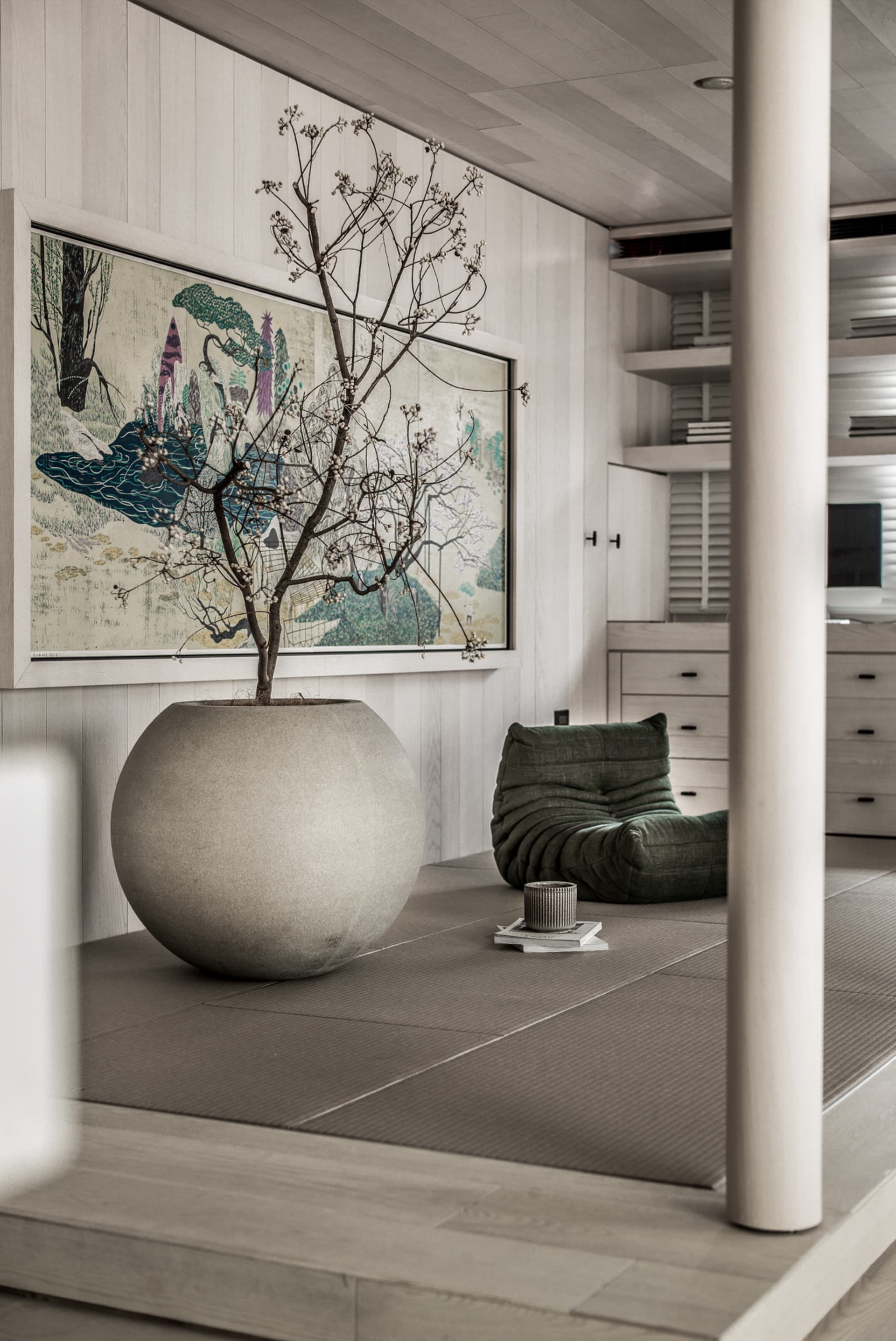 A serene living room with wooden floors, a large decorative pot with plants, and a minimalist green seat.