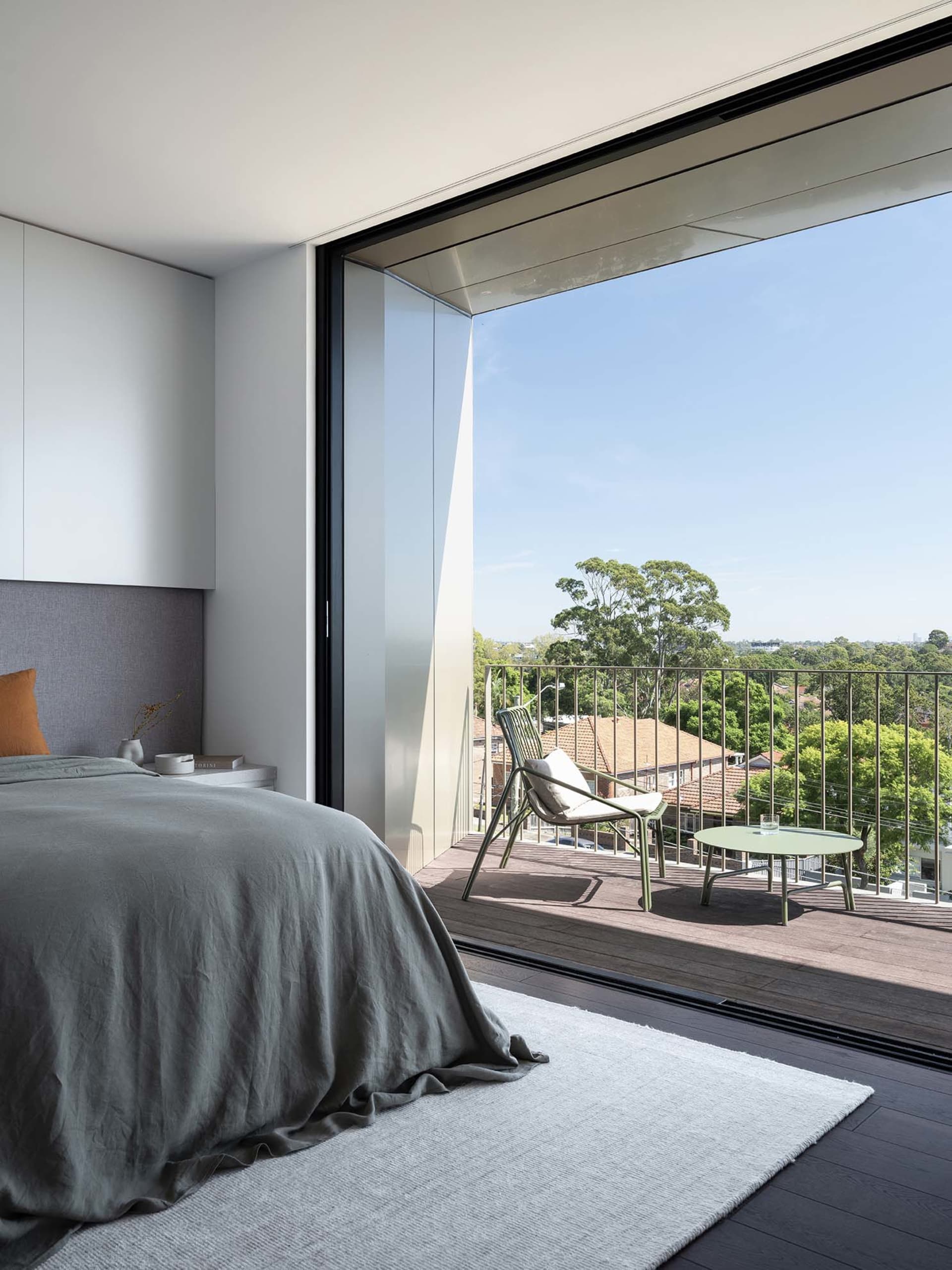 A modern bedroom with a wooden floor, featuring a bed, a rug, and a large window overlooking a balcony with a chair and table