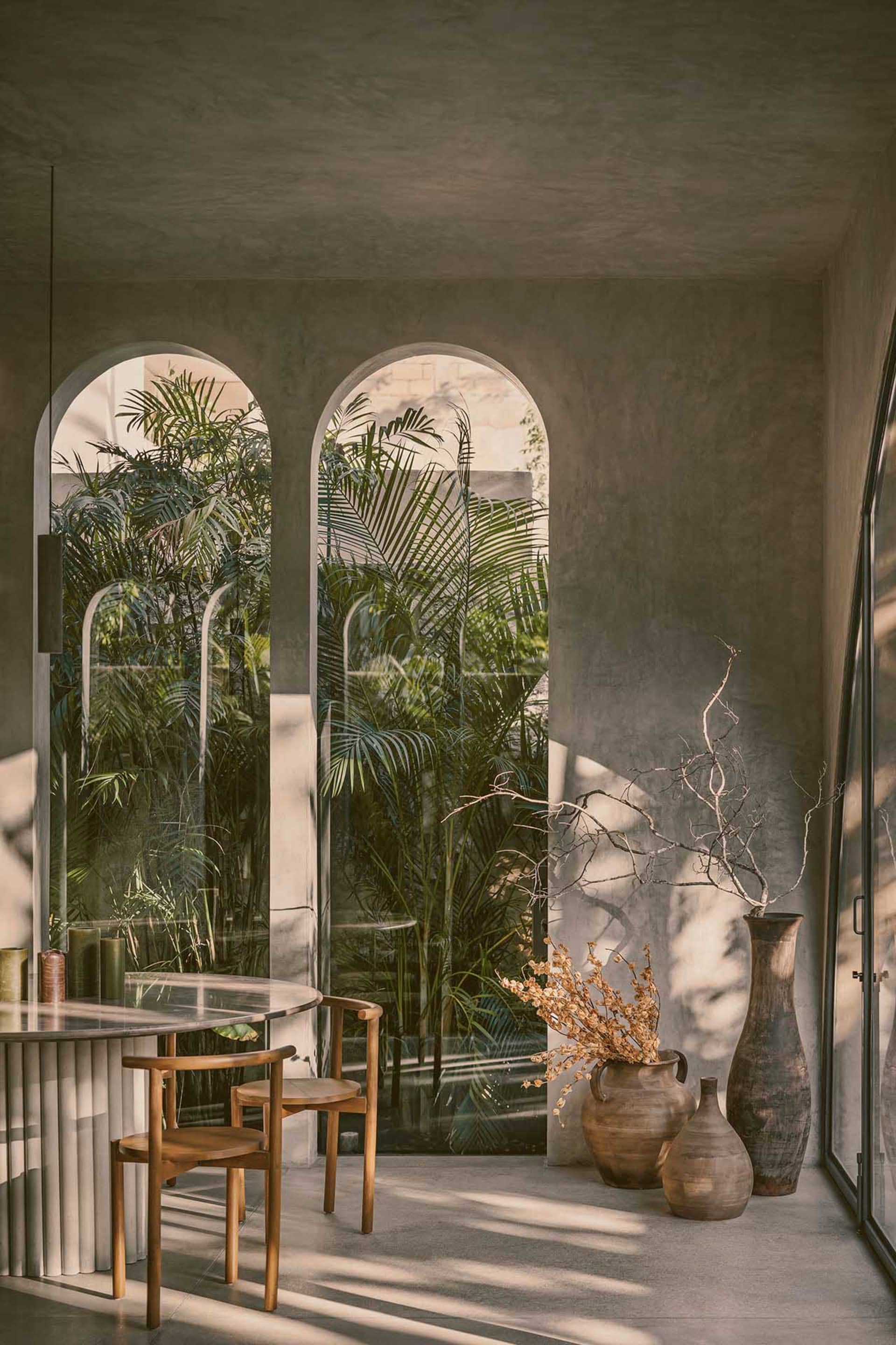 A modern, minimalist living room with concrete walls and floor, featuring a clear view of palm trees outside through arched w