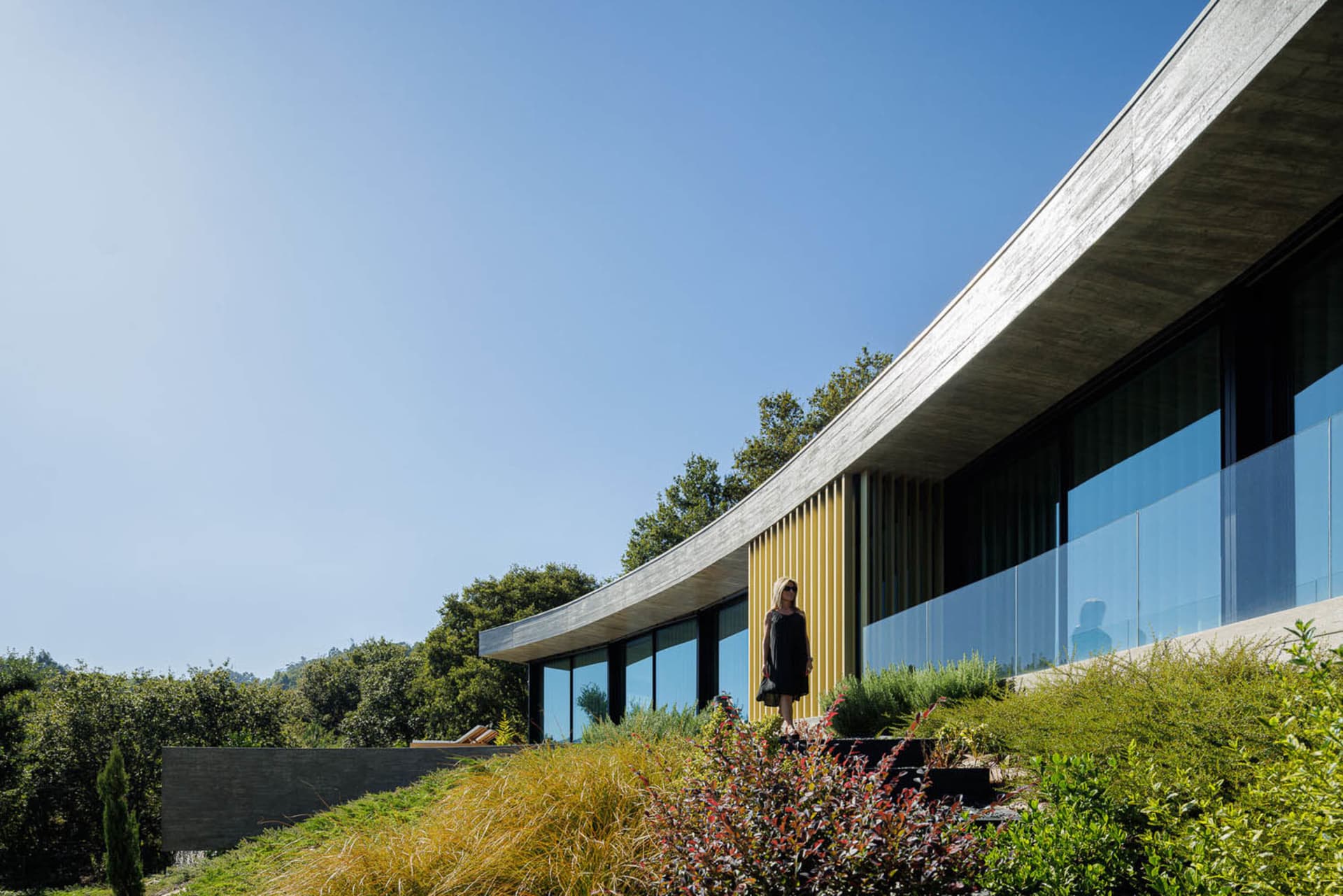 A modern house with a glass exterior and a sloped, curved roof set against a clear blue sky with lush greenery in the backgro