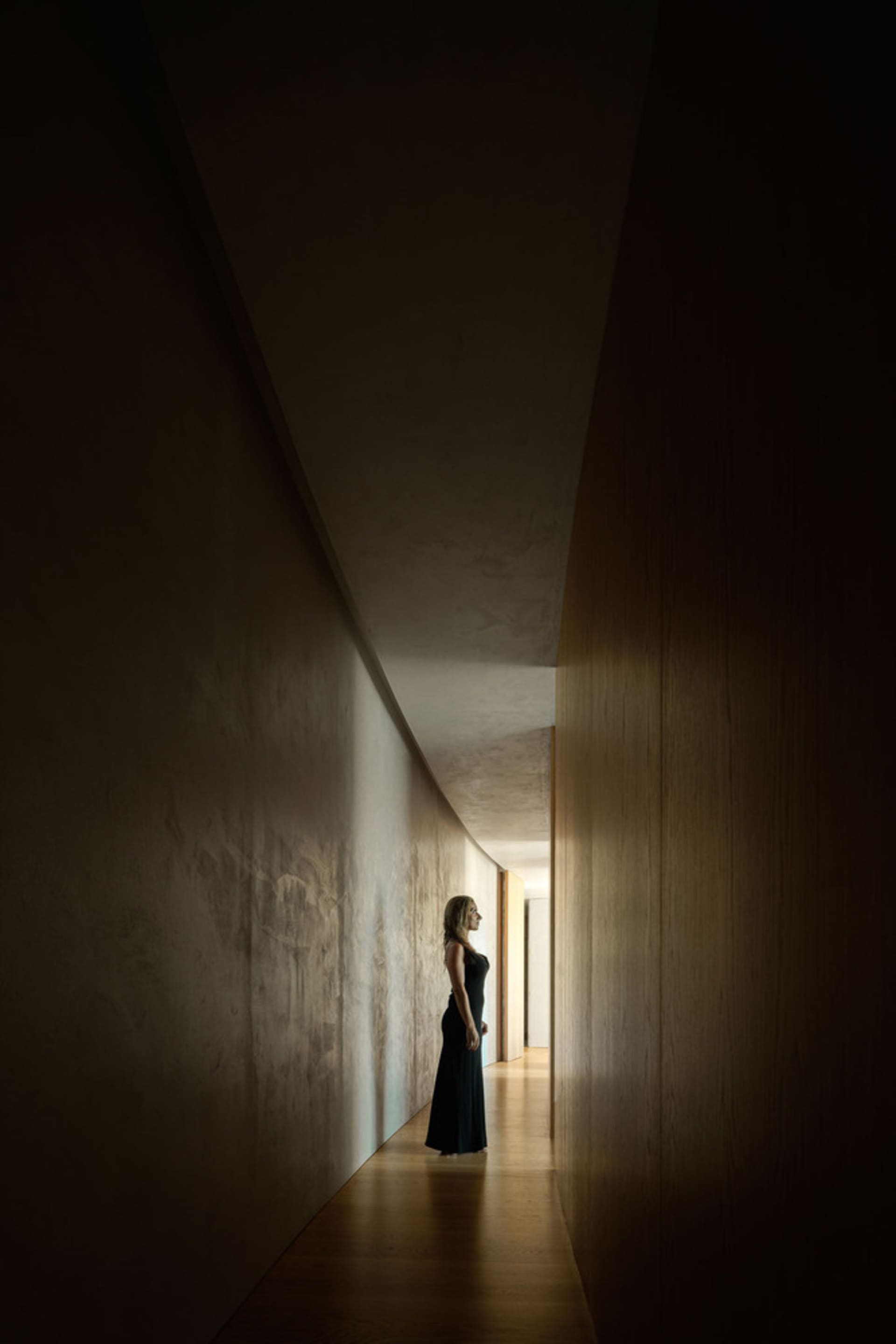 A woman in a black dress standing in a dark hallway with wooden walls and a gray light on the end.
