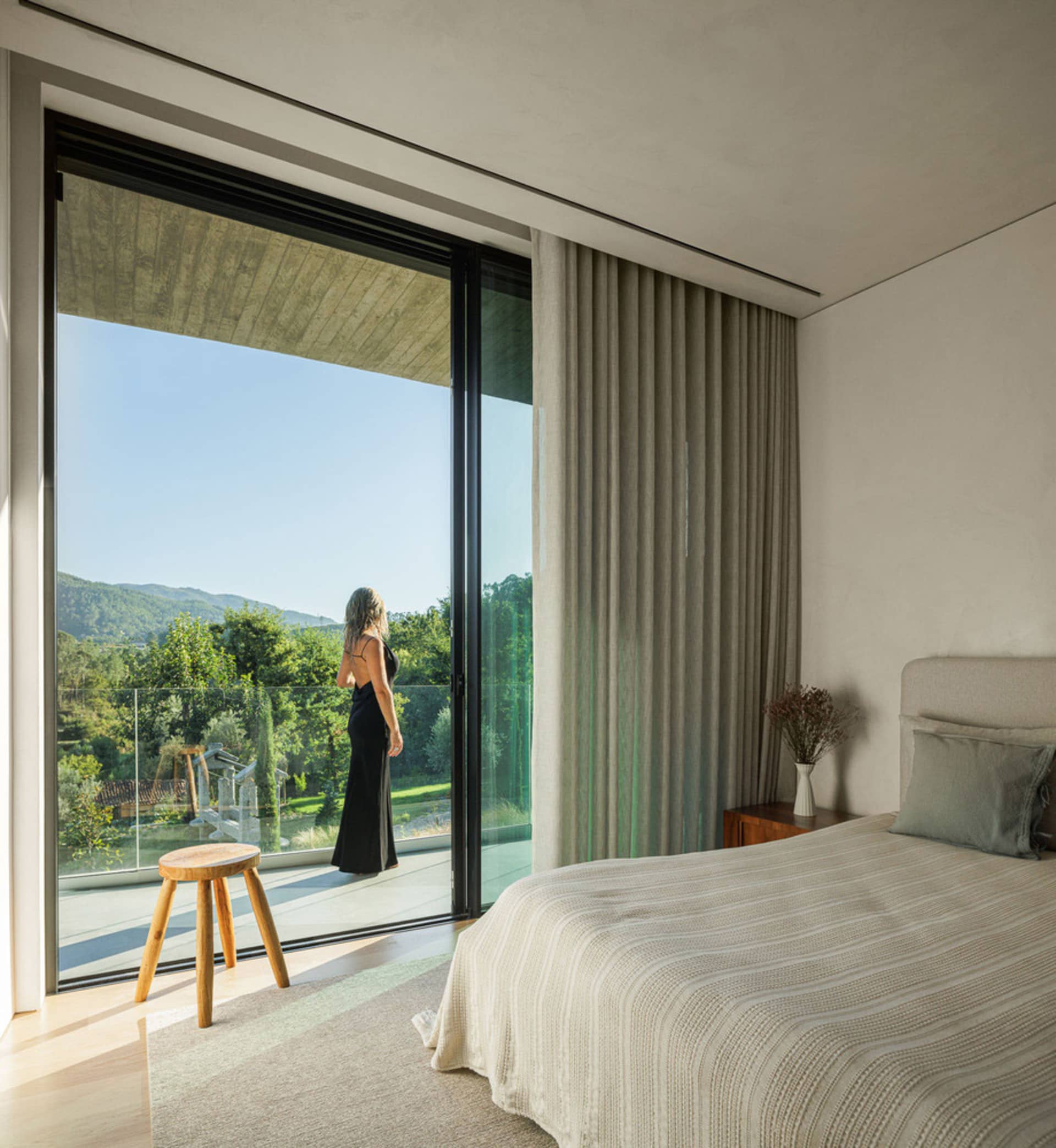 A modern bedroom with a glass door providing a view of greenery outside, featuring a canvas window treatment and a single woo
