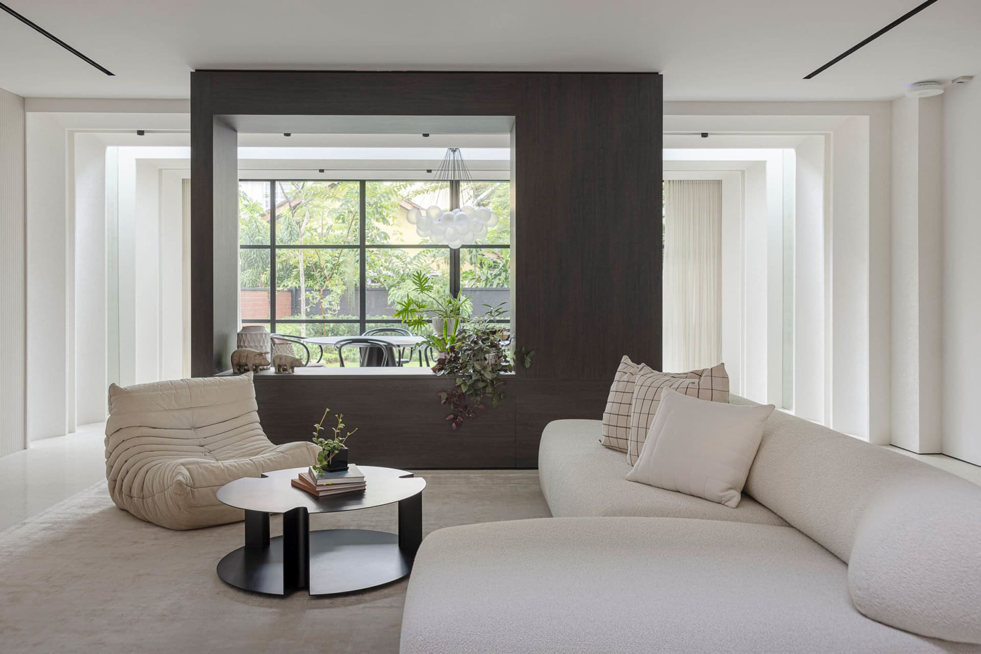 A modern living room with white soft textiles, minimalist furniture, and a view of the garden through large windows.