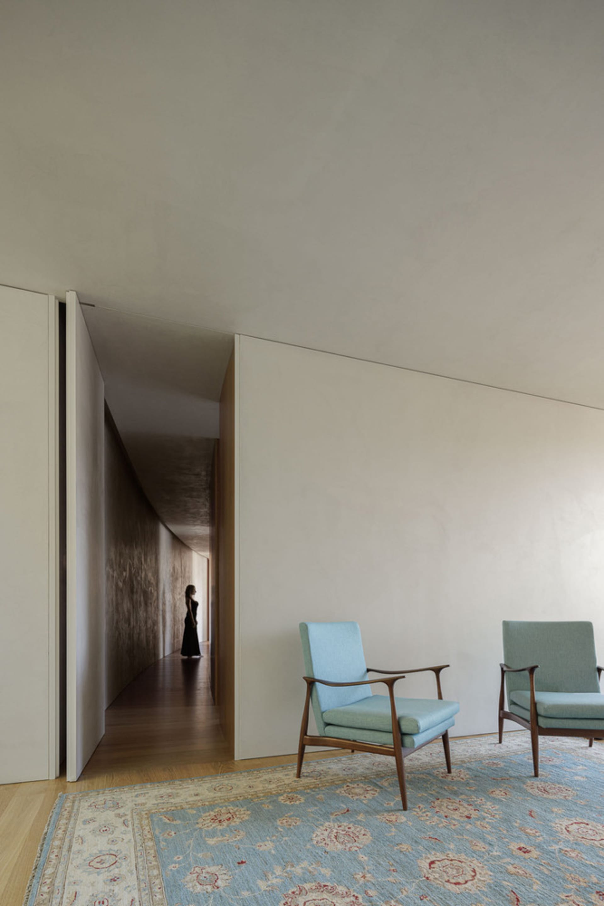 A minimalist living room with fabric-covered walls and wooden furniture, featuring a blue armchair on a floral-patterned rug.