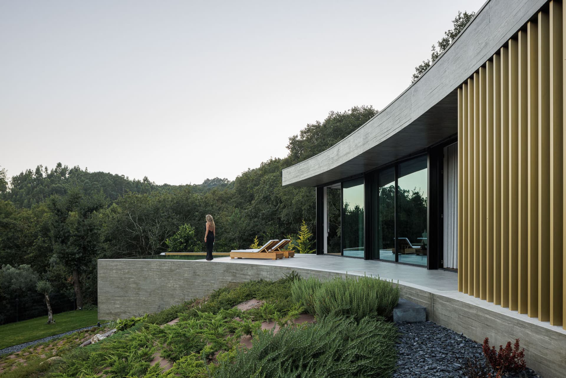 A modern residential exterior with concrete walls and large glass windows overlooking a forested area with two lounge chairs 