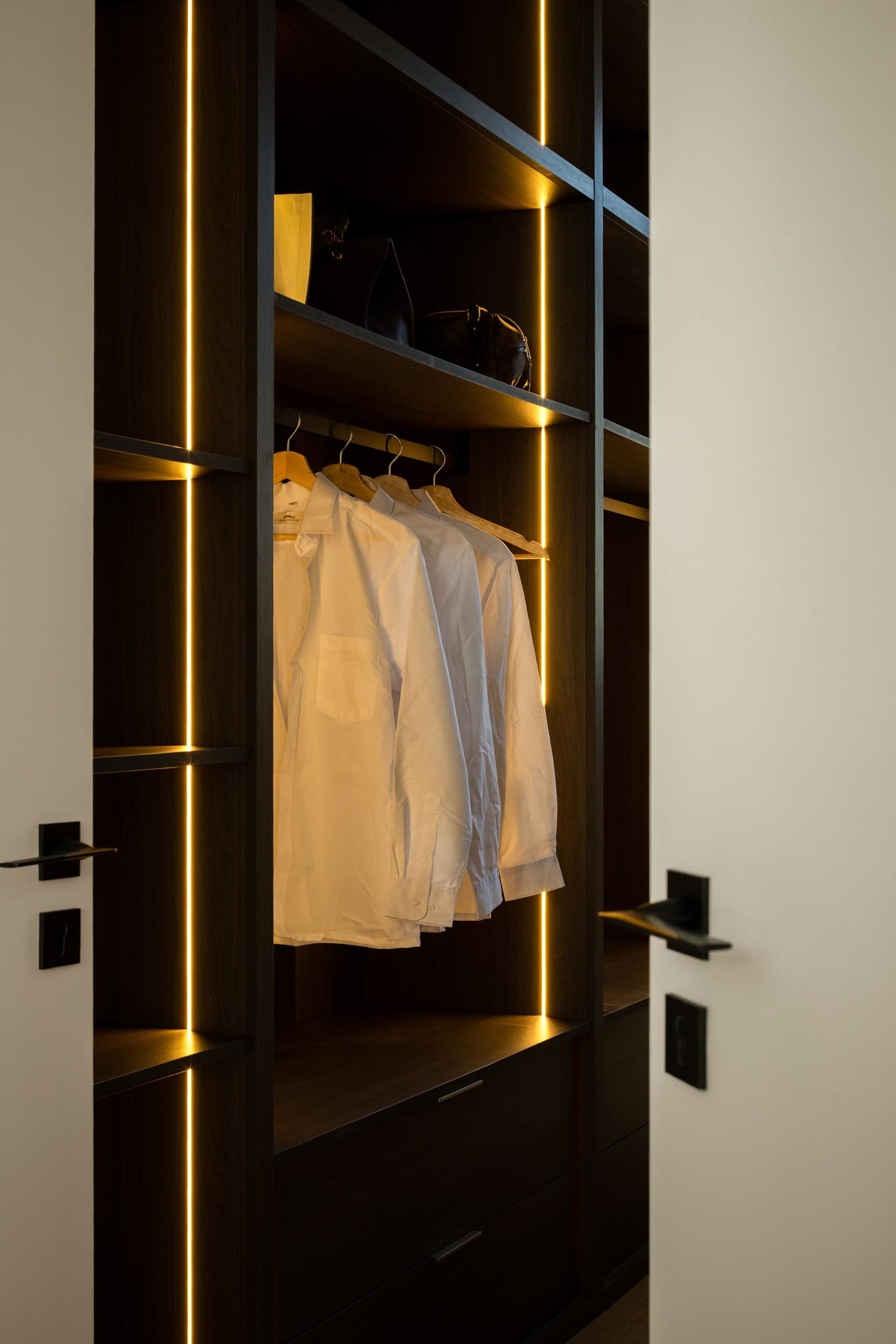 Modern wardrobe with white shirts on hangers and illuminated wooden structure with vertical lines, minimalist design.