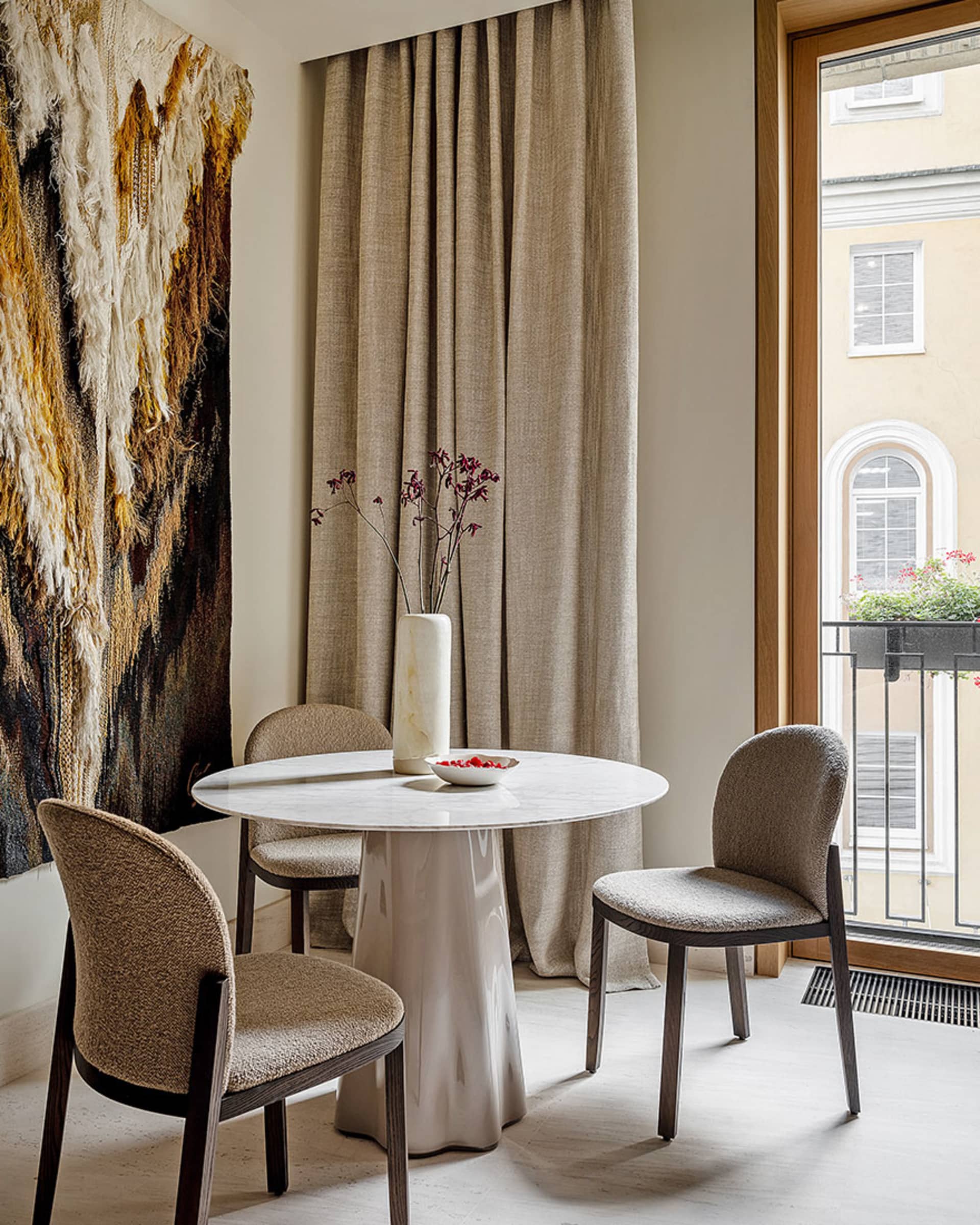 Modern dining room furnished with a round marble table and fabric chairs, featuring a decorative wall hanging and a vase with