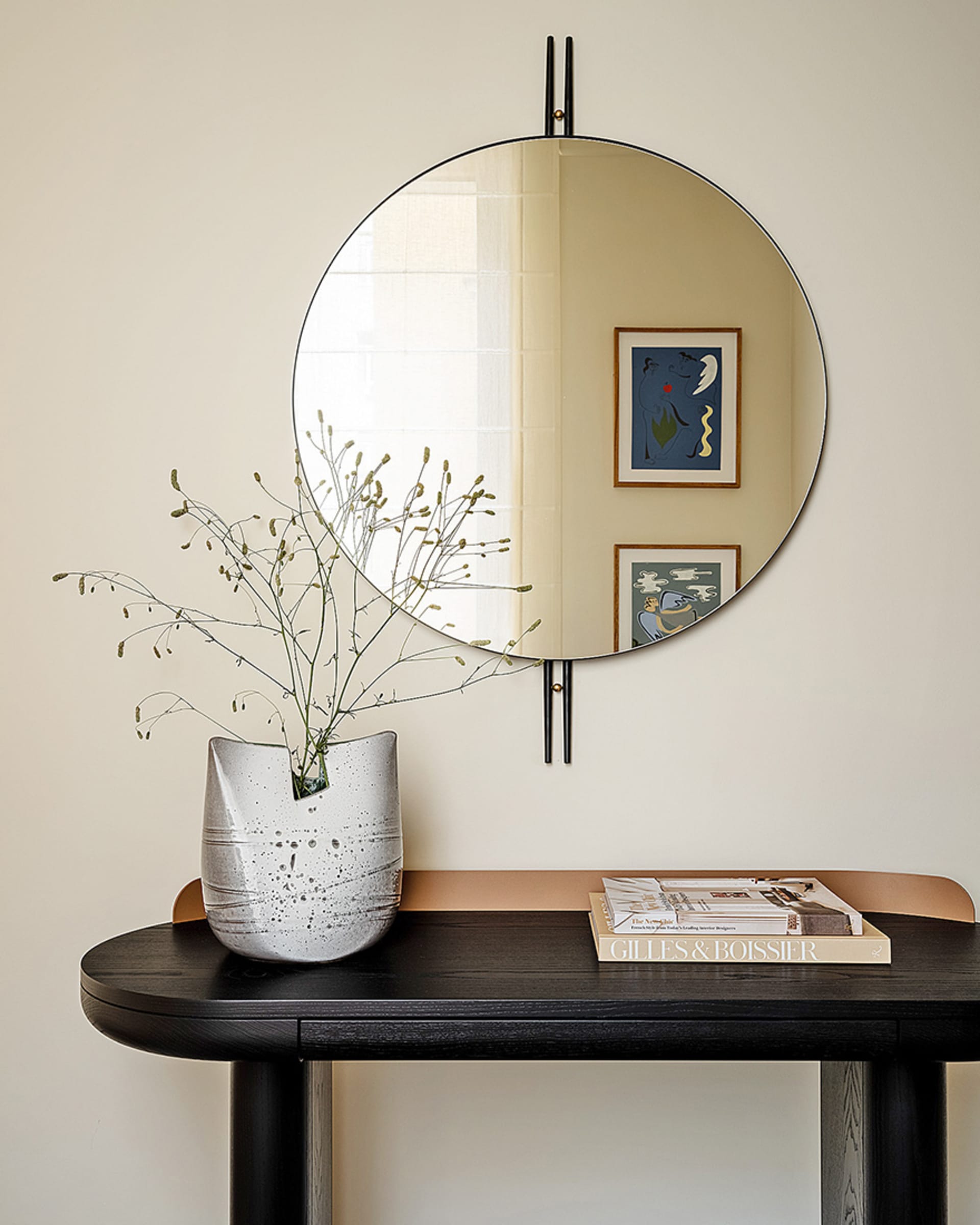 A modern interior design featuring a ceramic vase with dried flowers and a round mirror on the wall, books placed on a dark t