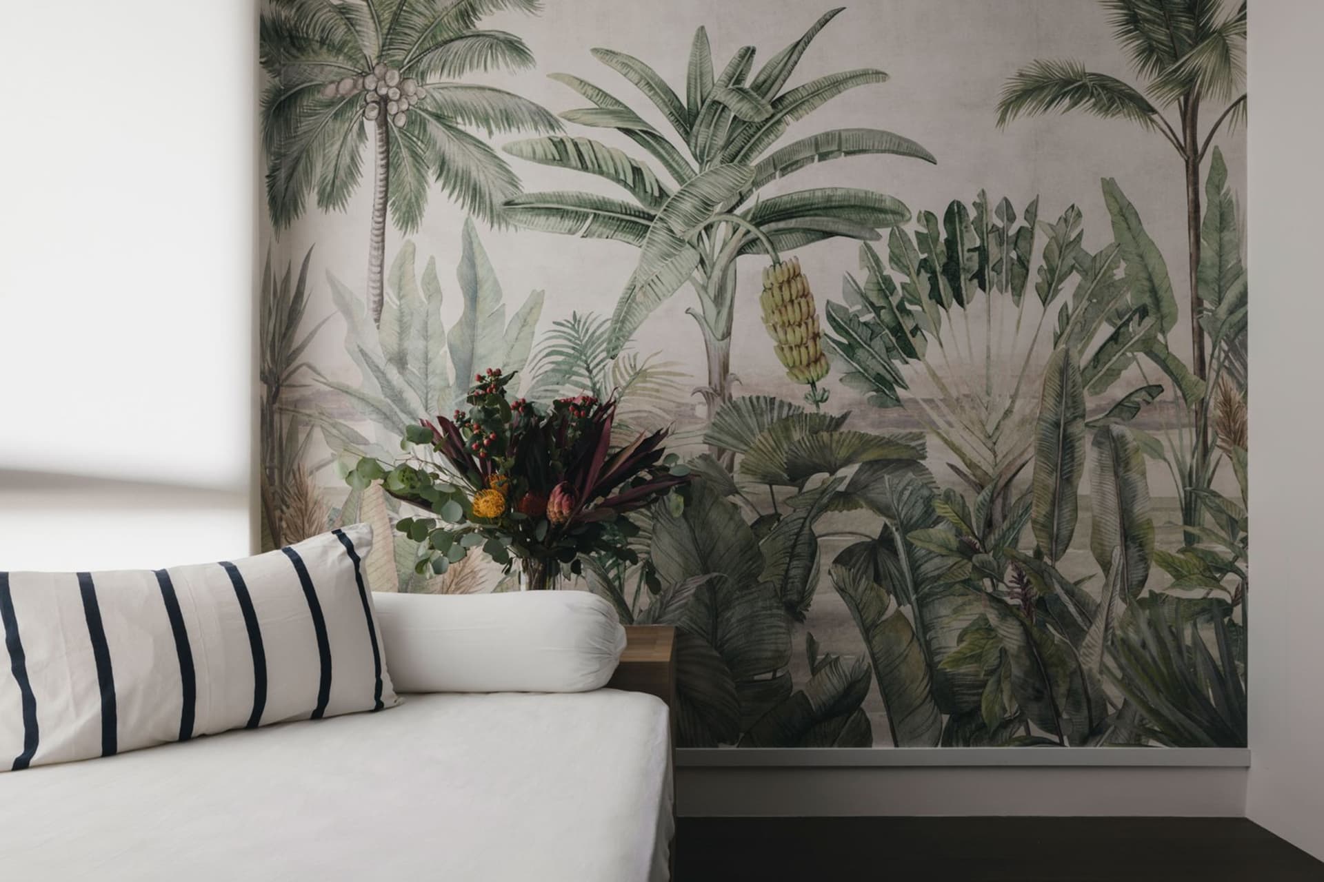 A cozy living space with a white sofa, striped pillows, and a floral arrangement in front of a botanical-themed wallpaper wal