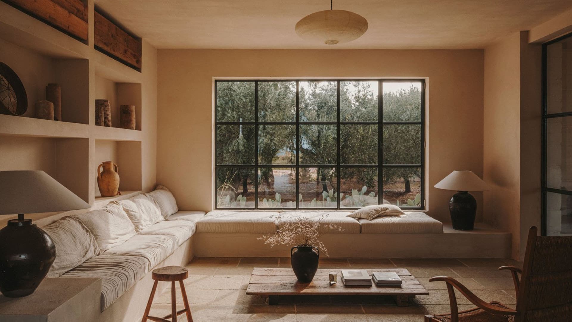 A minimalist living room featuring concrete walls, a large window overlooking an olive grove, and a wooden coffee table with