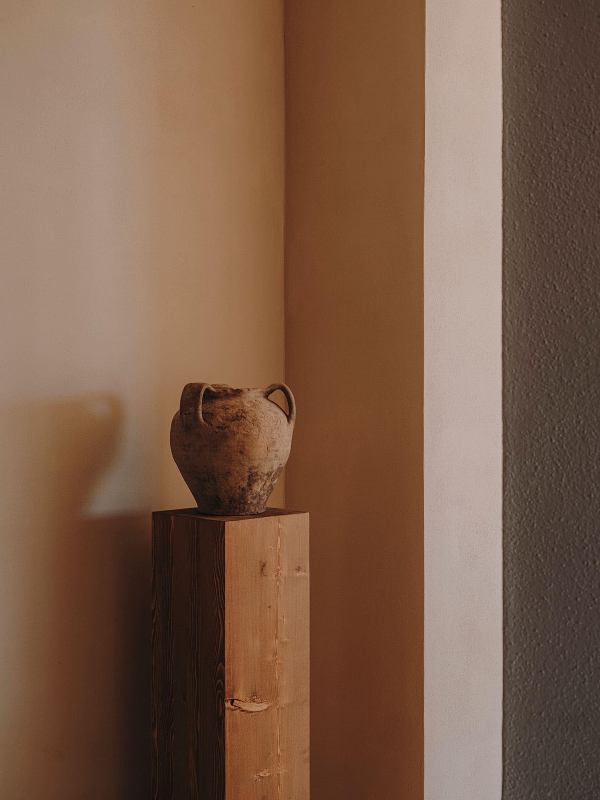 A minimalist ceramic vase with two handles placed on a wooden pedestal in a minimalist setting.