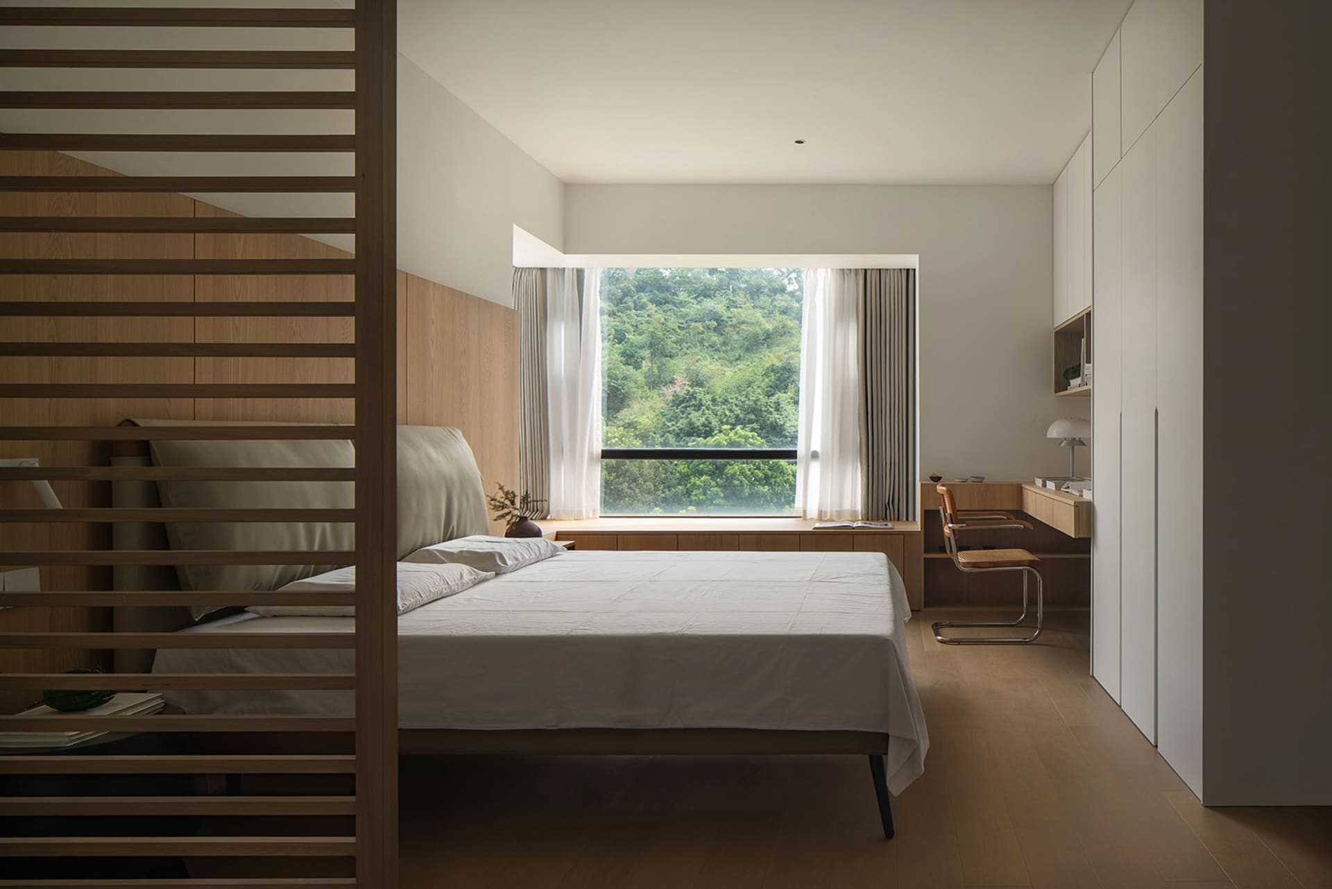 A minimalist bedroom featuring a bed with white sheets and a wooden headboard and floor, with a large window showcasing a lus