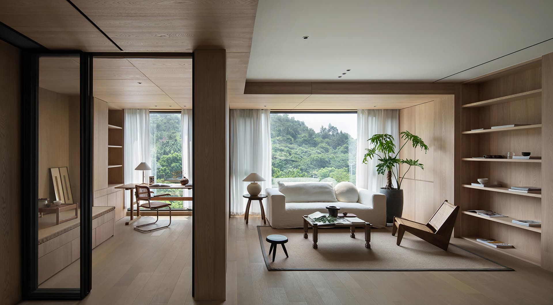A modern minimalist living room featuring a white couch, wooden accents, and glass windows overlooking a lush green landscape