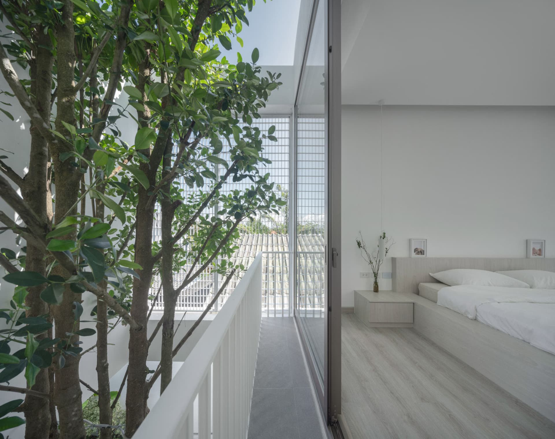 A minimalist bedroom with a white bed and wooden floor, featuring green trees outside the large windows and a clean, modern d